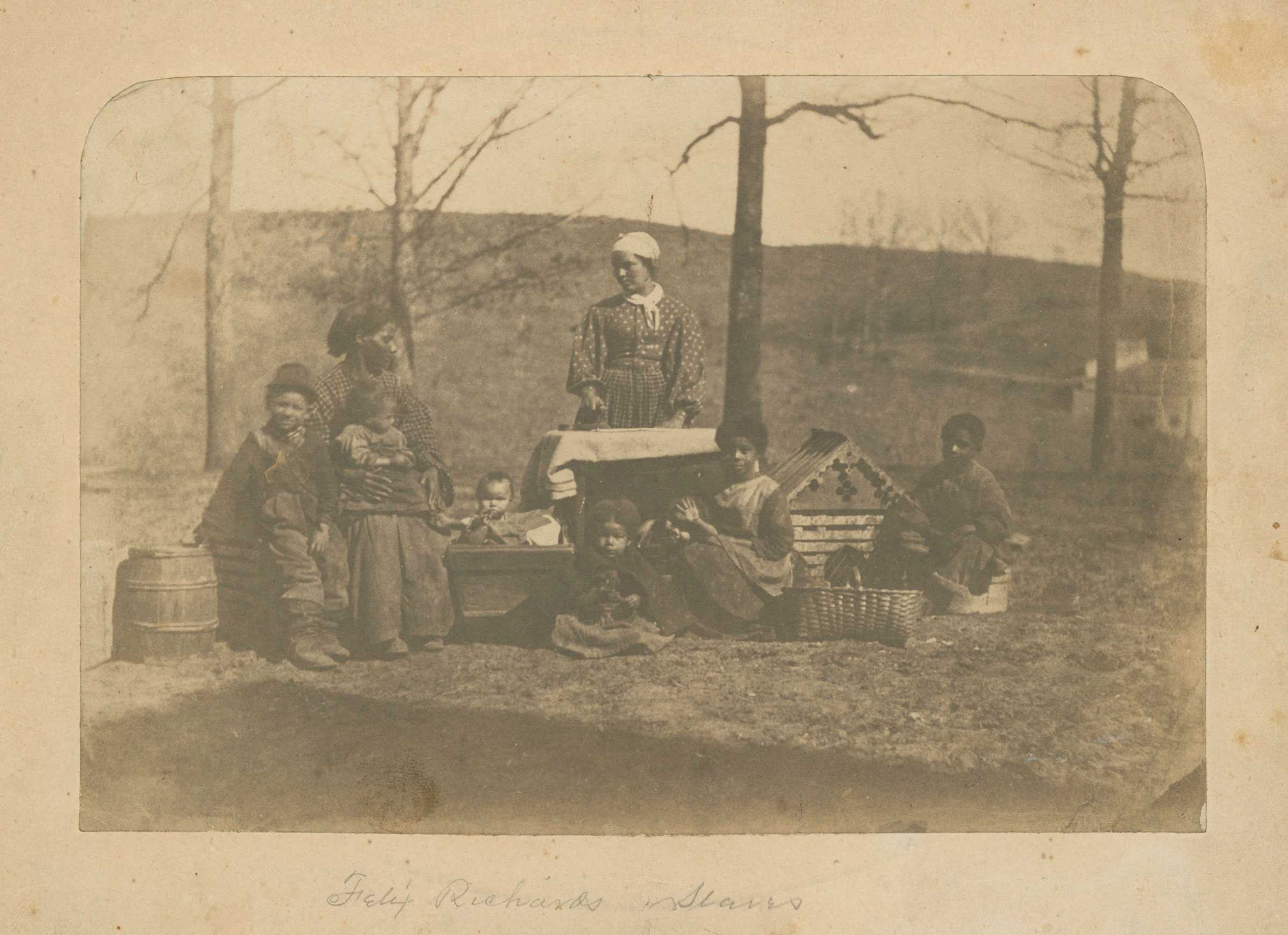 An albumen print depicting two enslaved women and seven children posed outside, with baskets and a cradle.