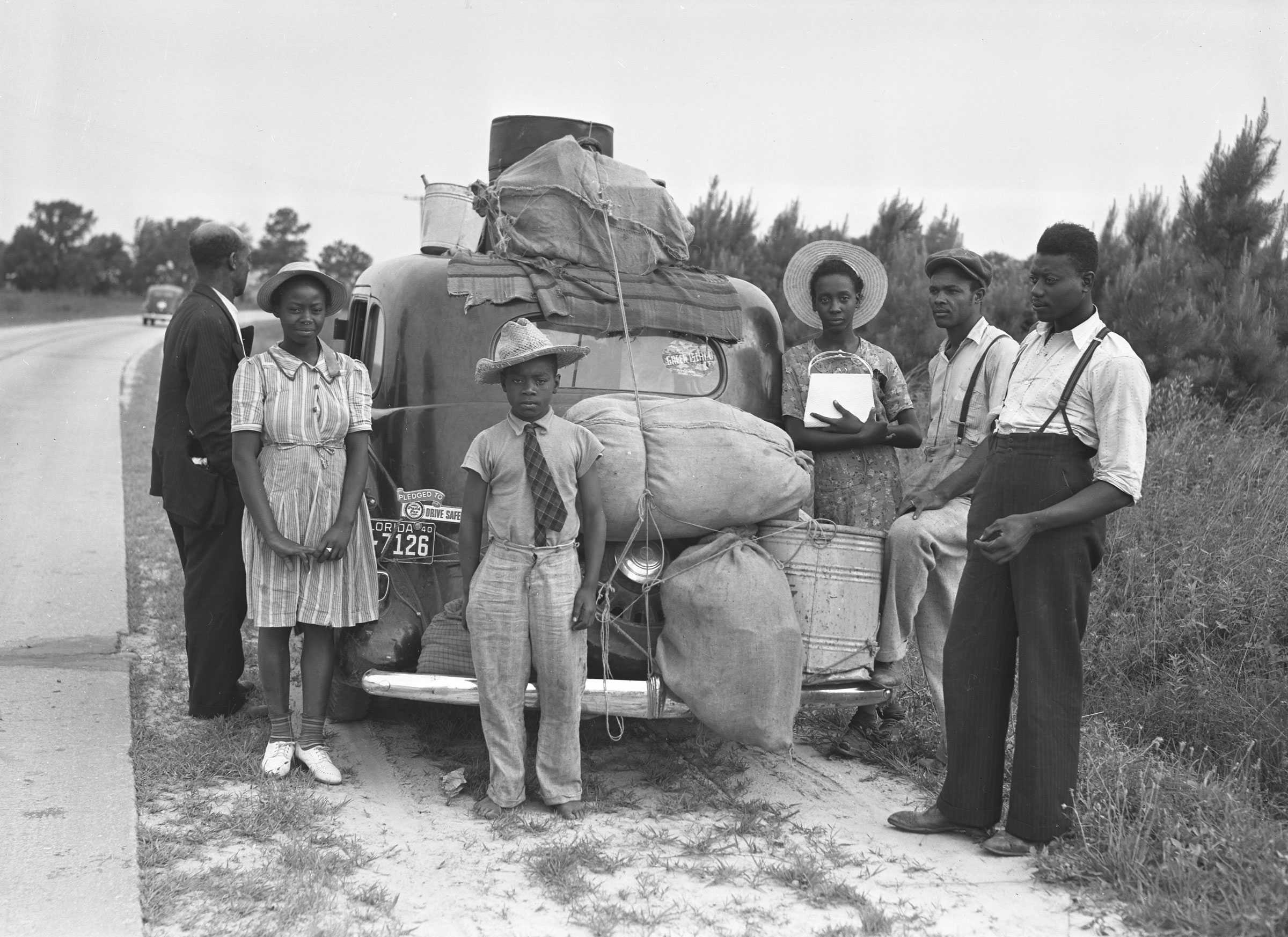 A black and white photograph of a family standing behind a packed car on the side of a road.