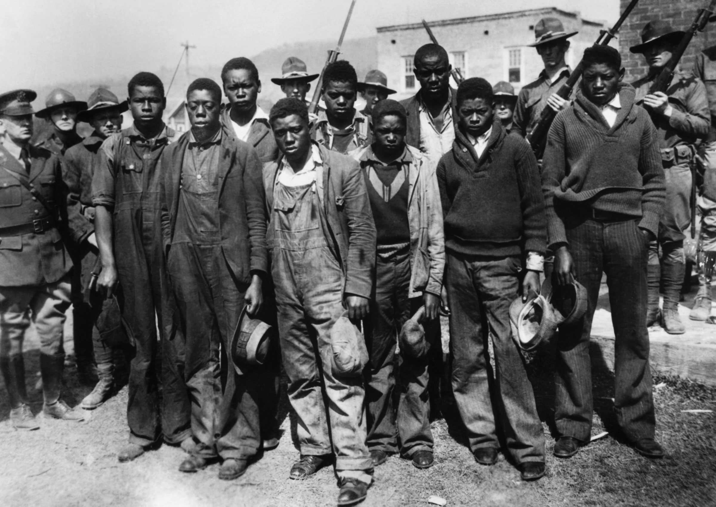 Black and white photograph showing a group of African American men standing in a group as men in uniforms guard them with rifles raised.