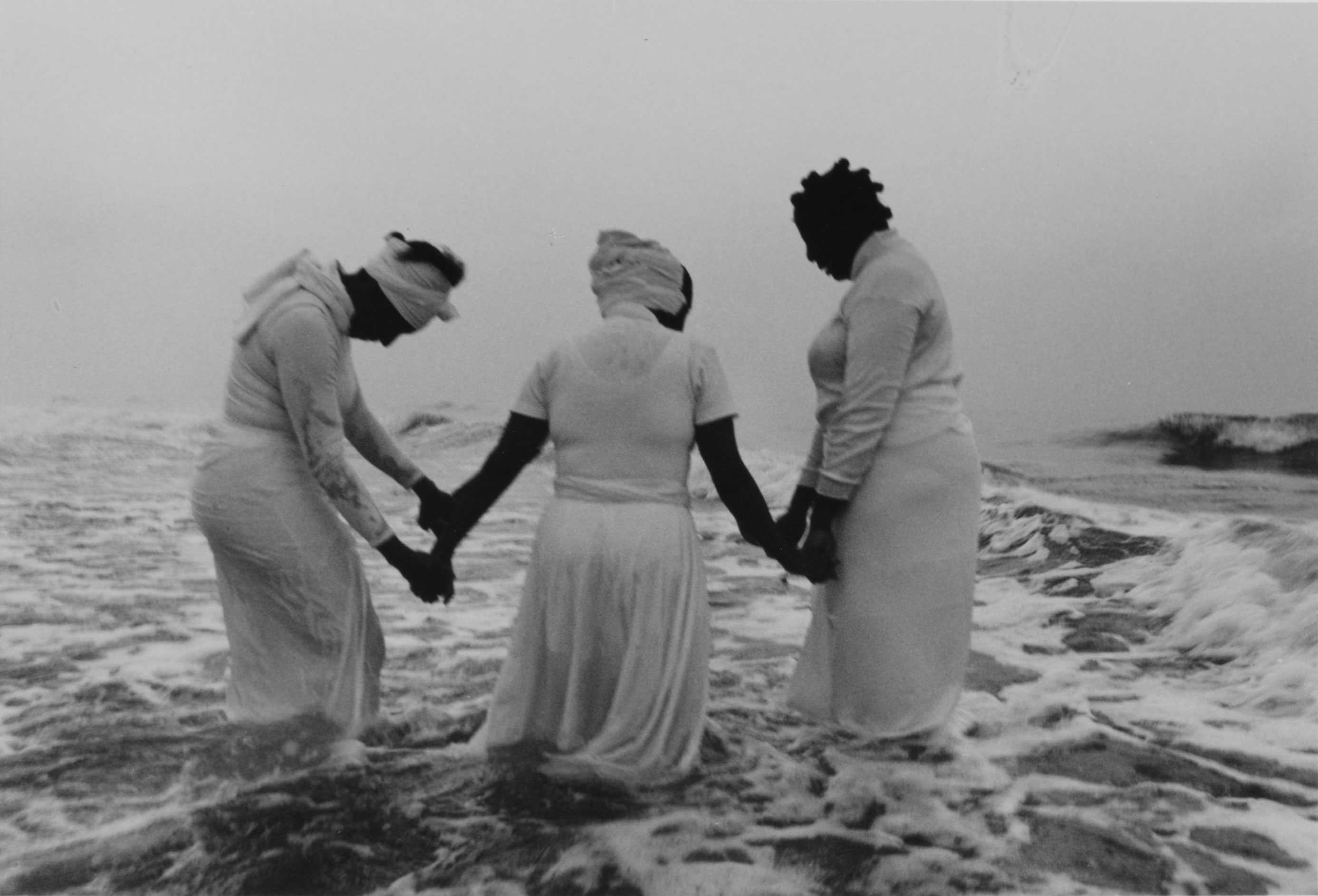 A black-and-white photo of three women in white dresses standing in ocean waves during a memorial ritual.