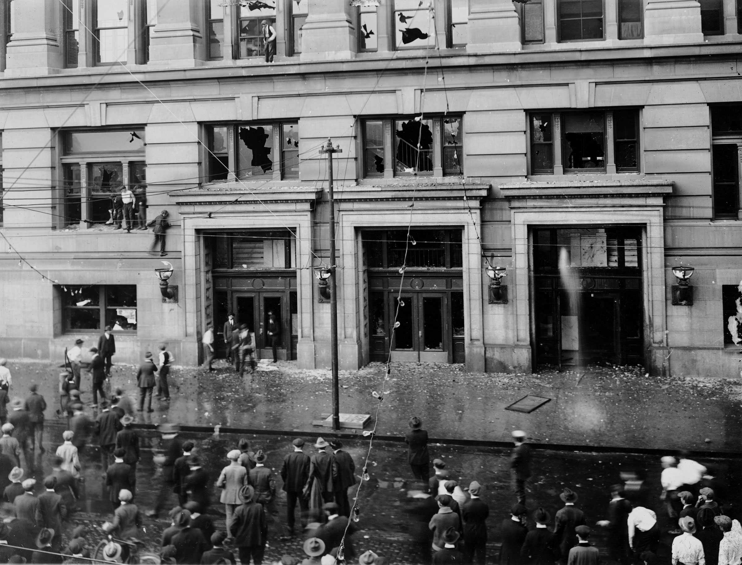 Black and white photograph showing building with rioters in and in front of building.  Glass is shattered everywhere.  The rioters are perched on window sills and in the street.