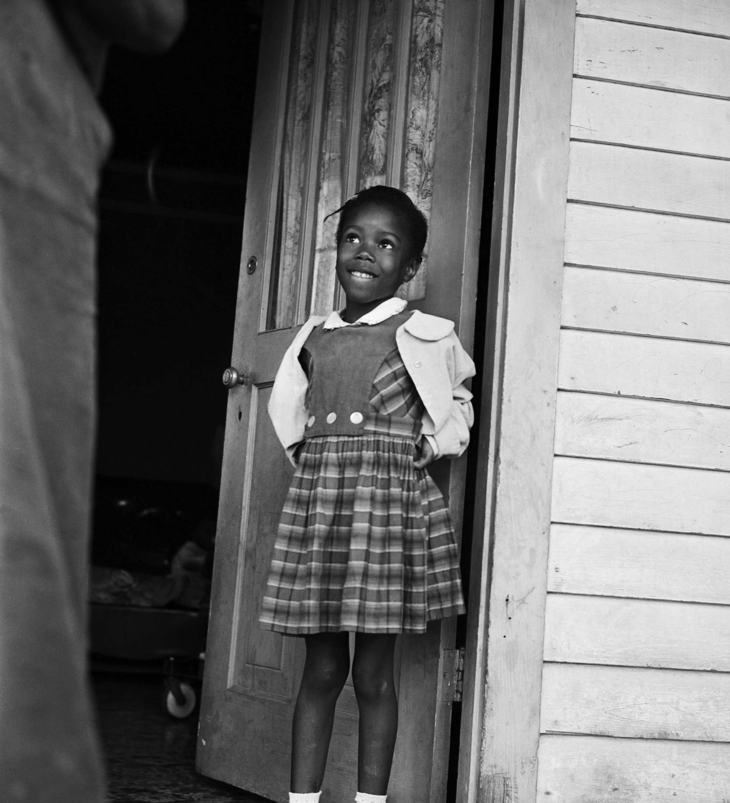 A black and white photograph of Ruby Bridges smiling up at an adult as she stands in a door frame.