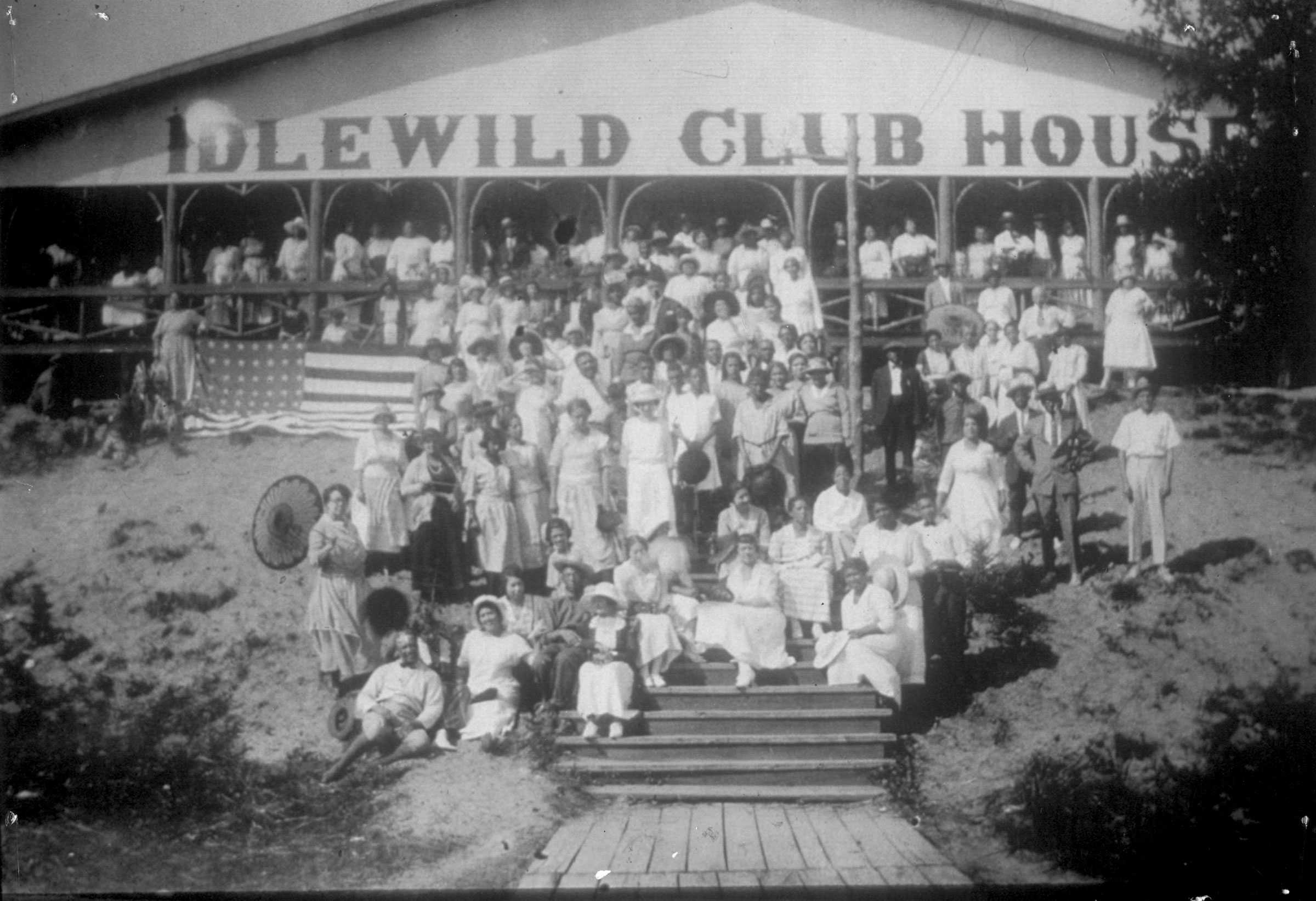 Black and white photograph showing large group of men and women posed before a structure identified as the"IDLEWILD CLUB HOUSE." Subjects are dressed mainly in light colored leisure wear.  There is a large American flag in the background.