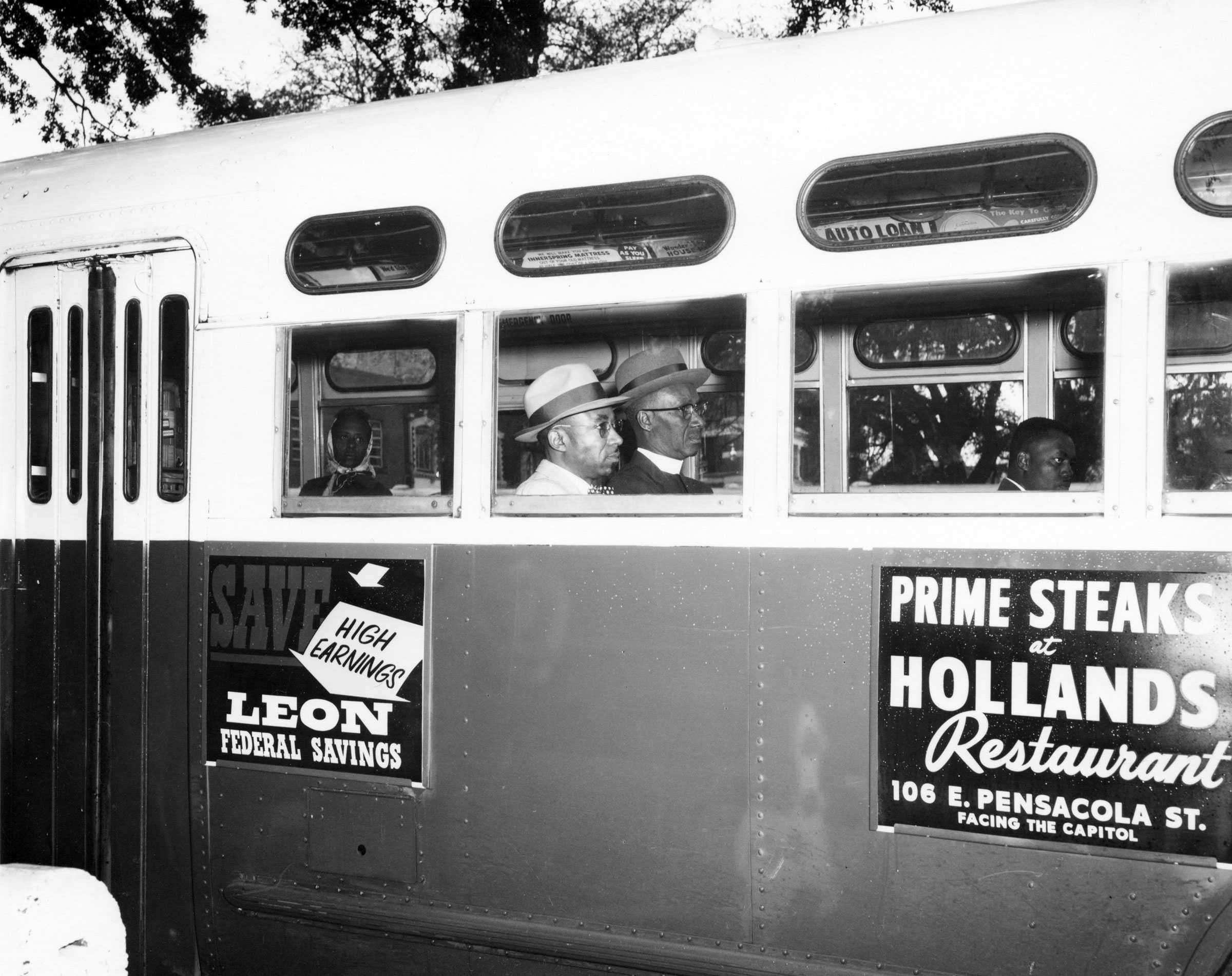 A black and white photograph of a bus in Tallahassee. The viewer can see through the windows that there are 3 men sitting on the bus.