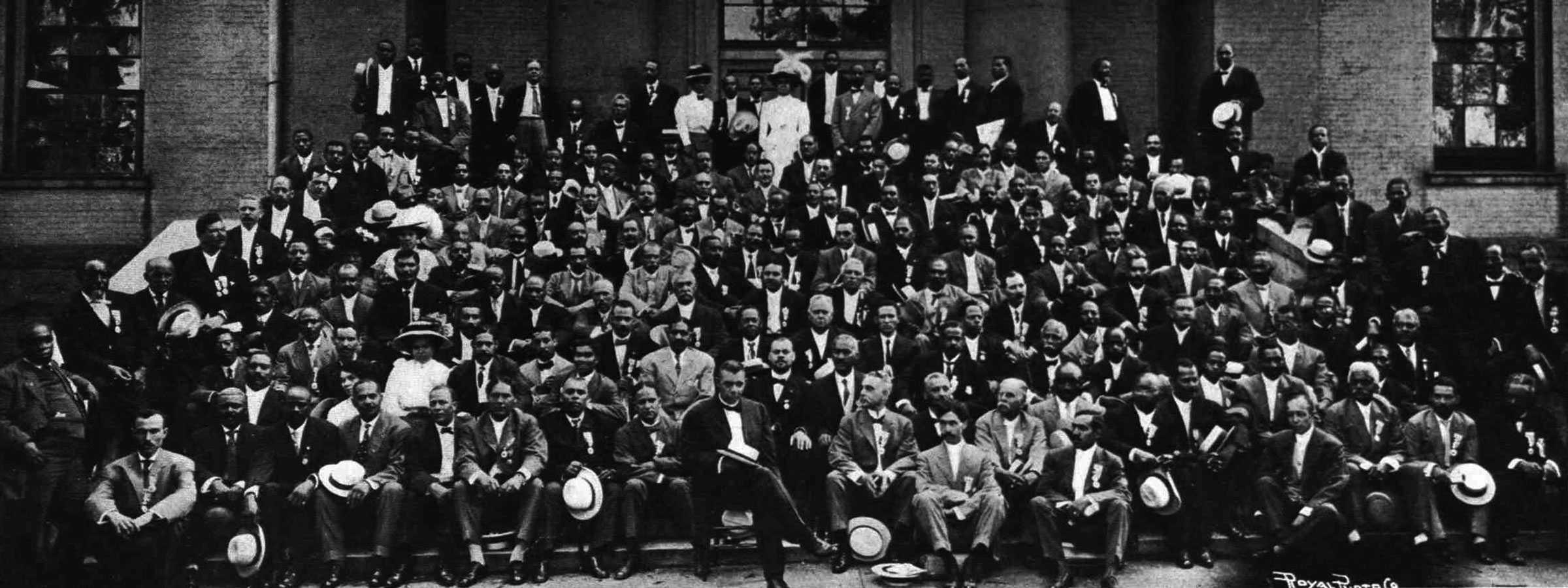 Large group portrait of men seated on the steps of a brick building.  There are a few women scattered in the crowd .