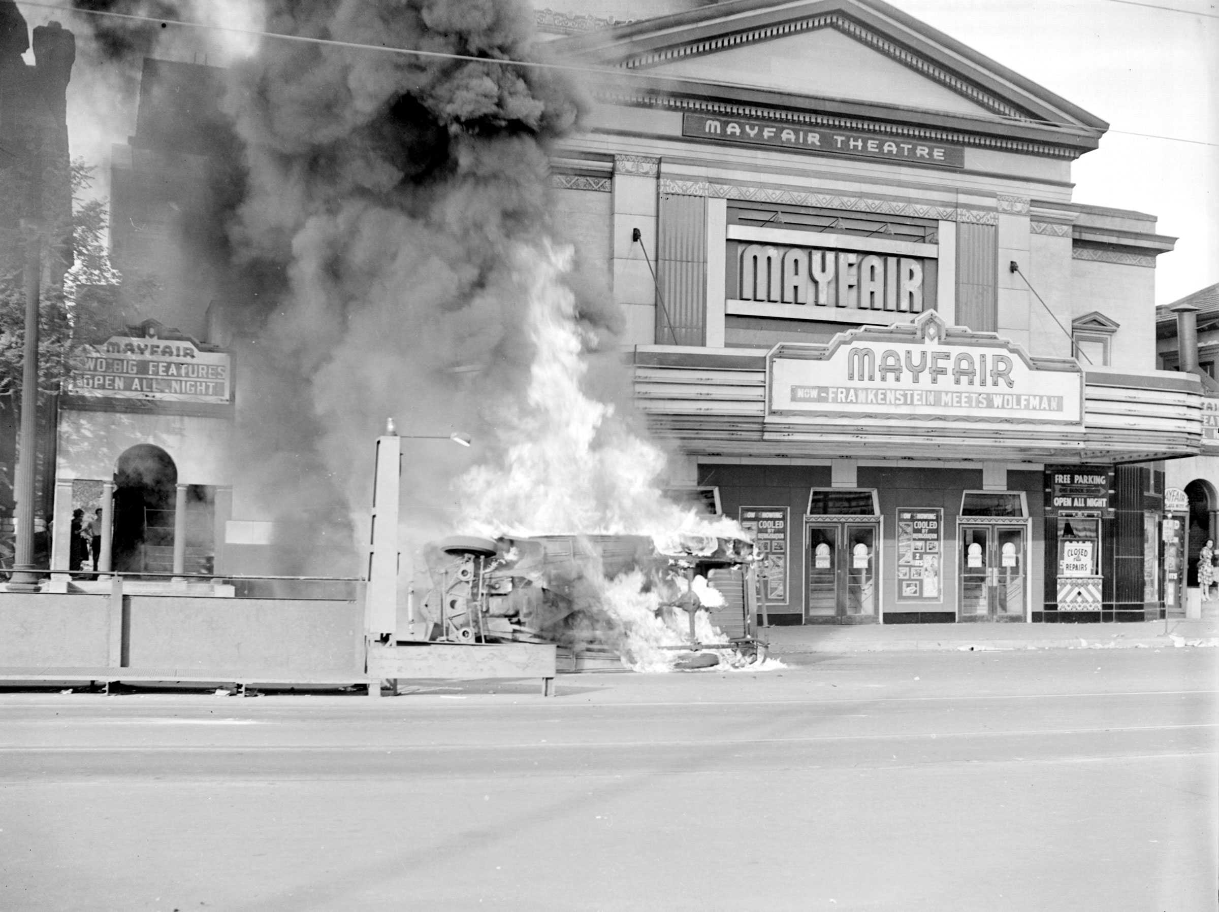 Black and white photograph showing the exterior of the"Mayfair Theatre" with a burning car on its side.