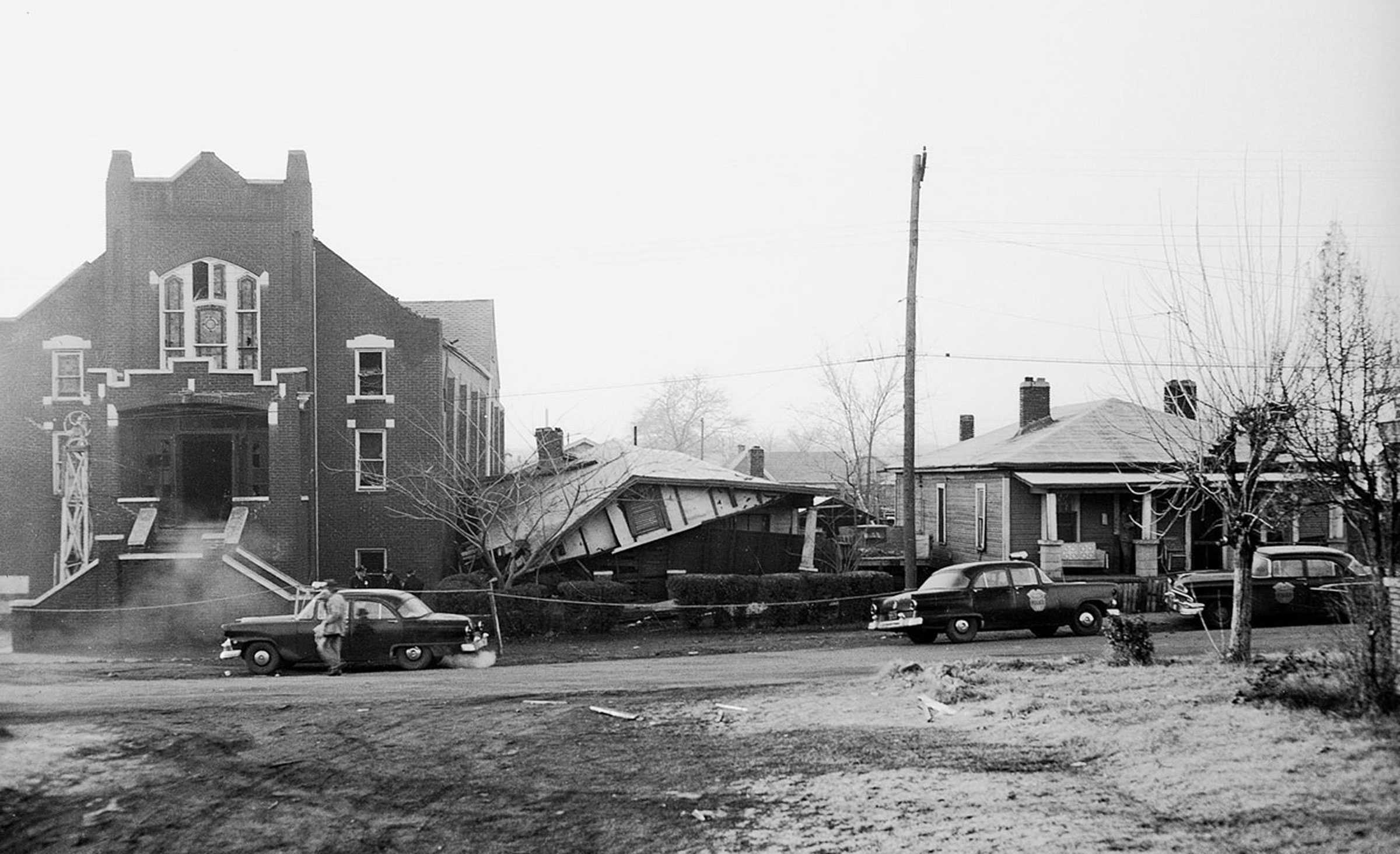 A black and white photograph of Bethel Baptist Church and the collapsed Shuttlesworth's home. Police officers and police cars line the street.