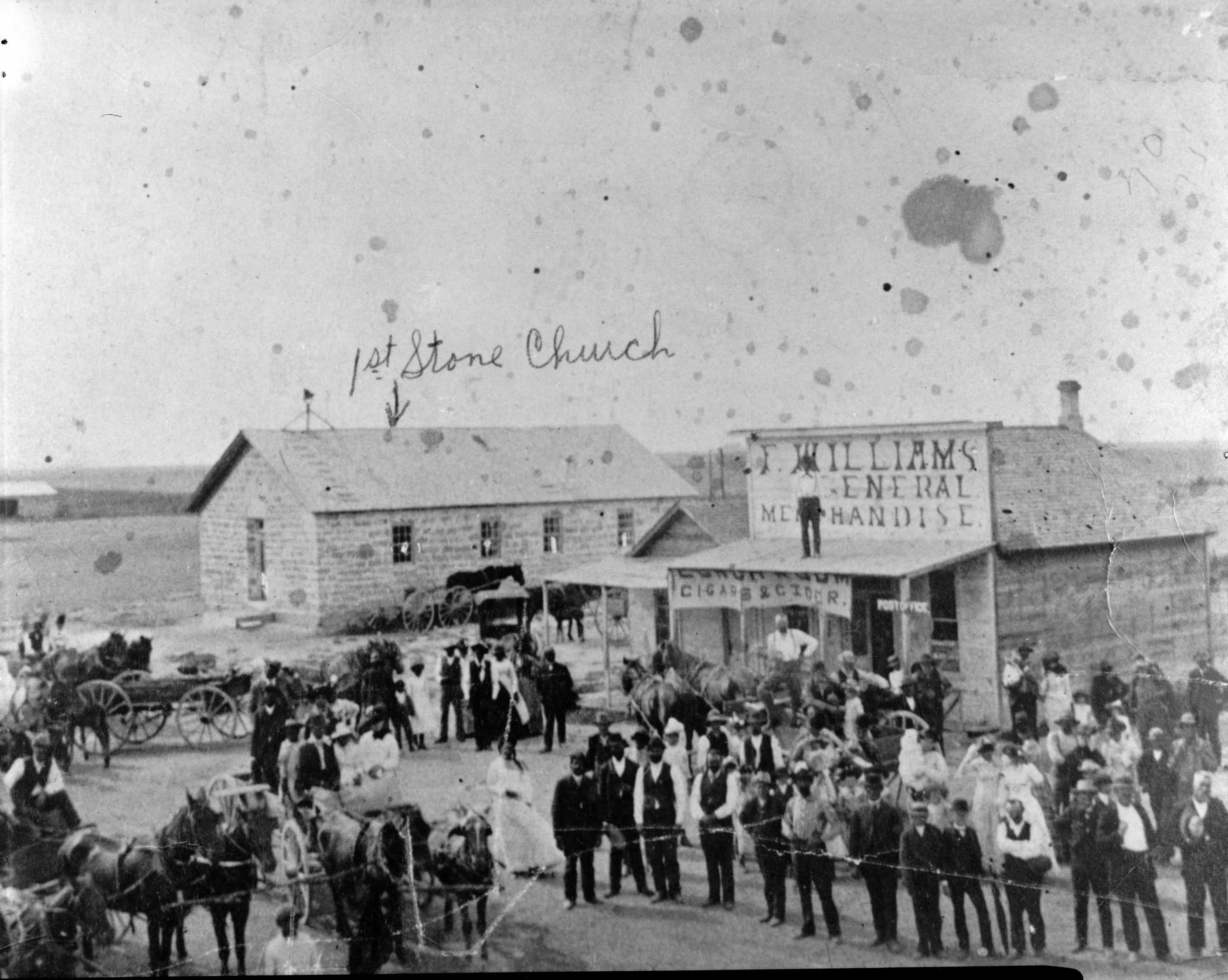 A worn black and white photograph of a rural town. A handwritten label and arrow pointing to the 1st Stone Church.