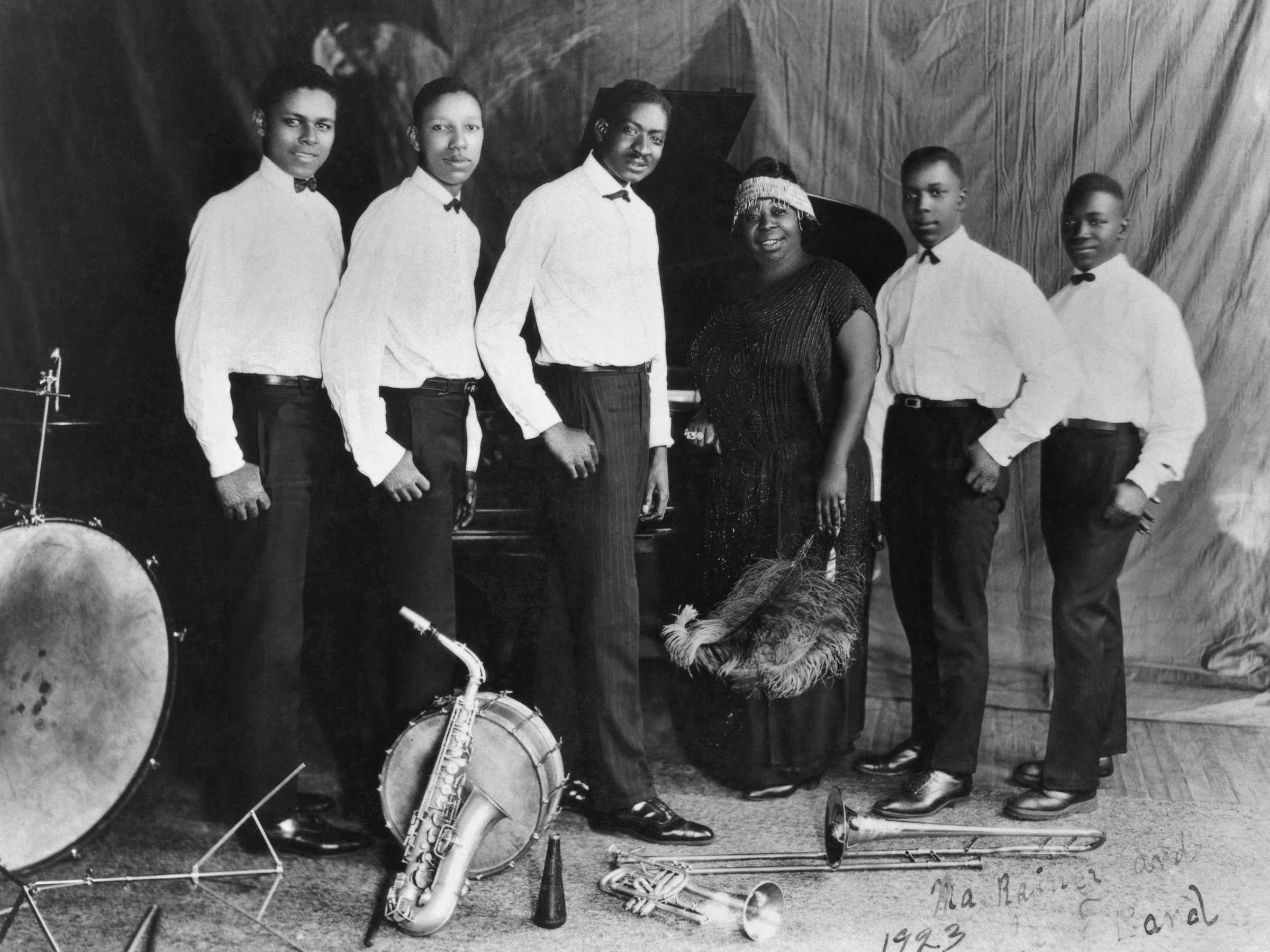 Group portrait of 5 male musicians wearing white shirts with bow ties and dark pants and a woman dressed in a long dark dress holding feathers.  There are various instruments playing at their feet including saxophone, trombone, trumpet and drums. There is writing in the bottom right hand corner of the photograph "Ma Rainey and / Band / 1923."