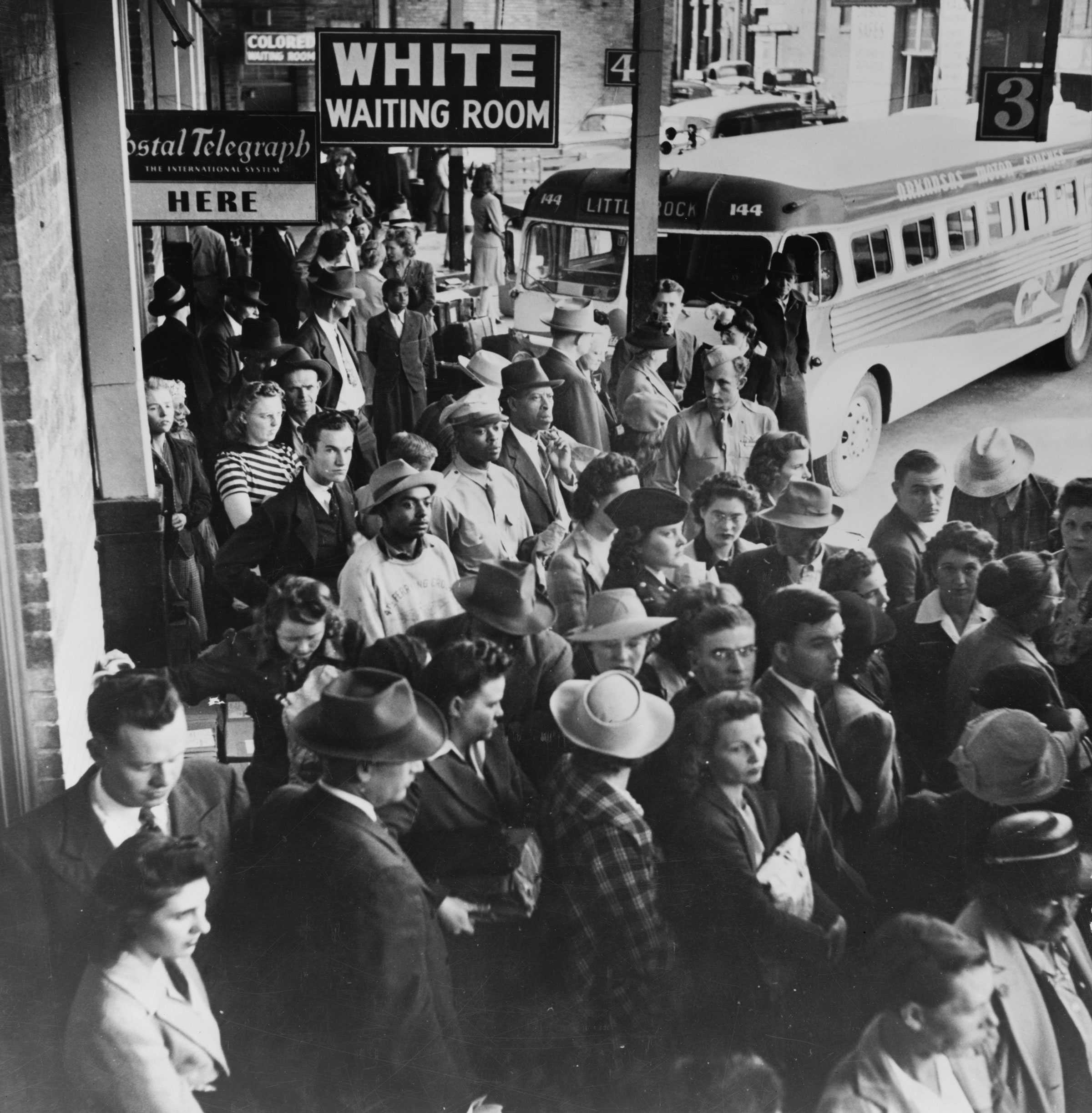 Black and white photograph of people waiting for a bus at a station.  There are buses visible in the background and a Sign that says "WHITE WAITING ROOM."