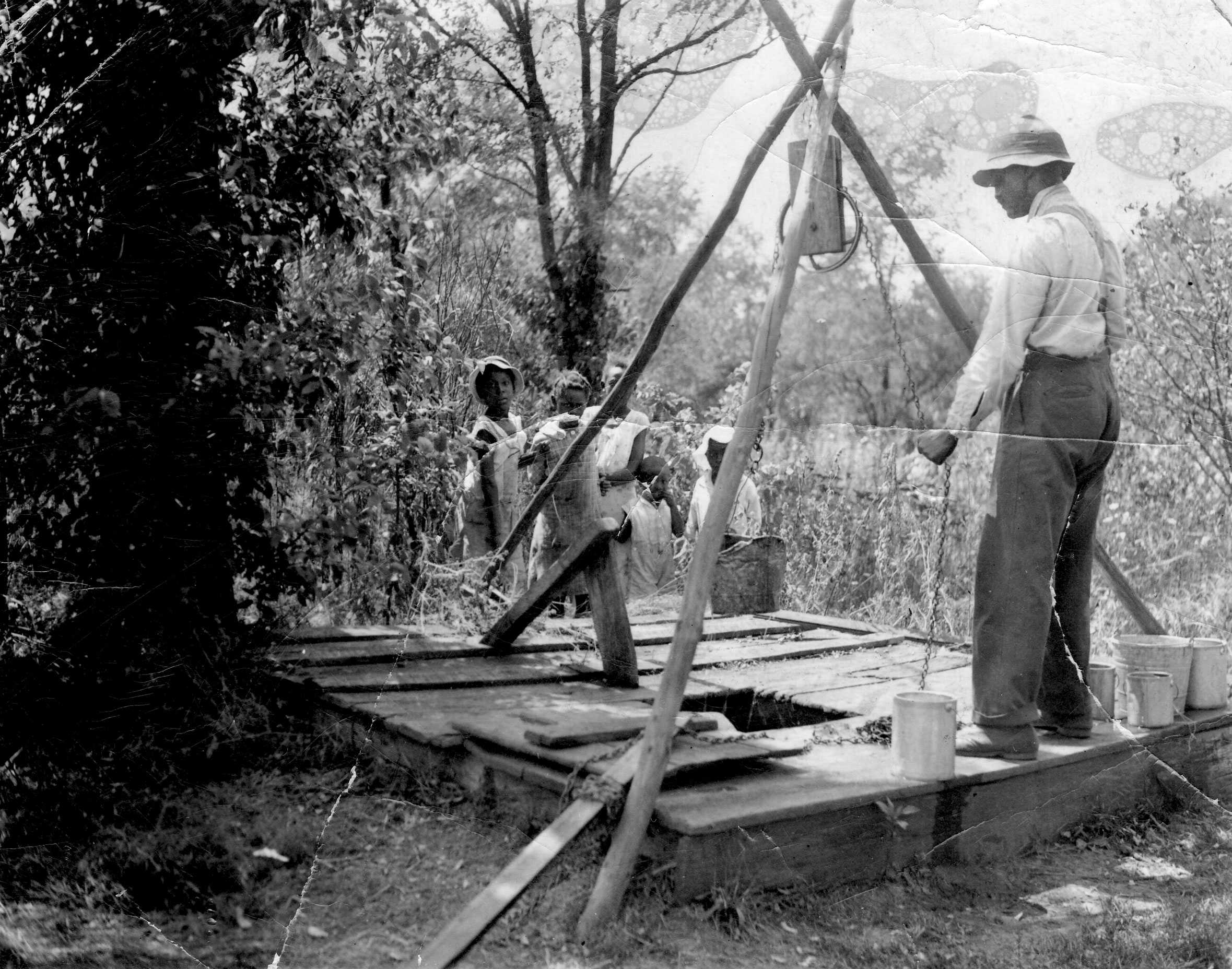 A worn photo of a family using a well. Kids are standing behind the well, while the father is pulling on the chain.