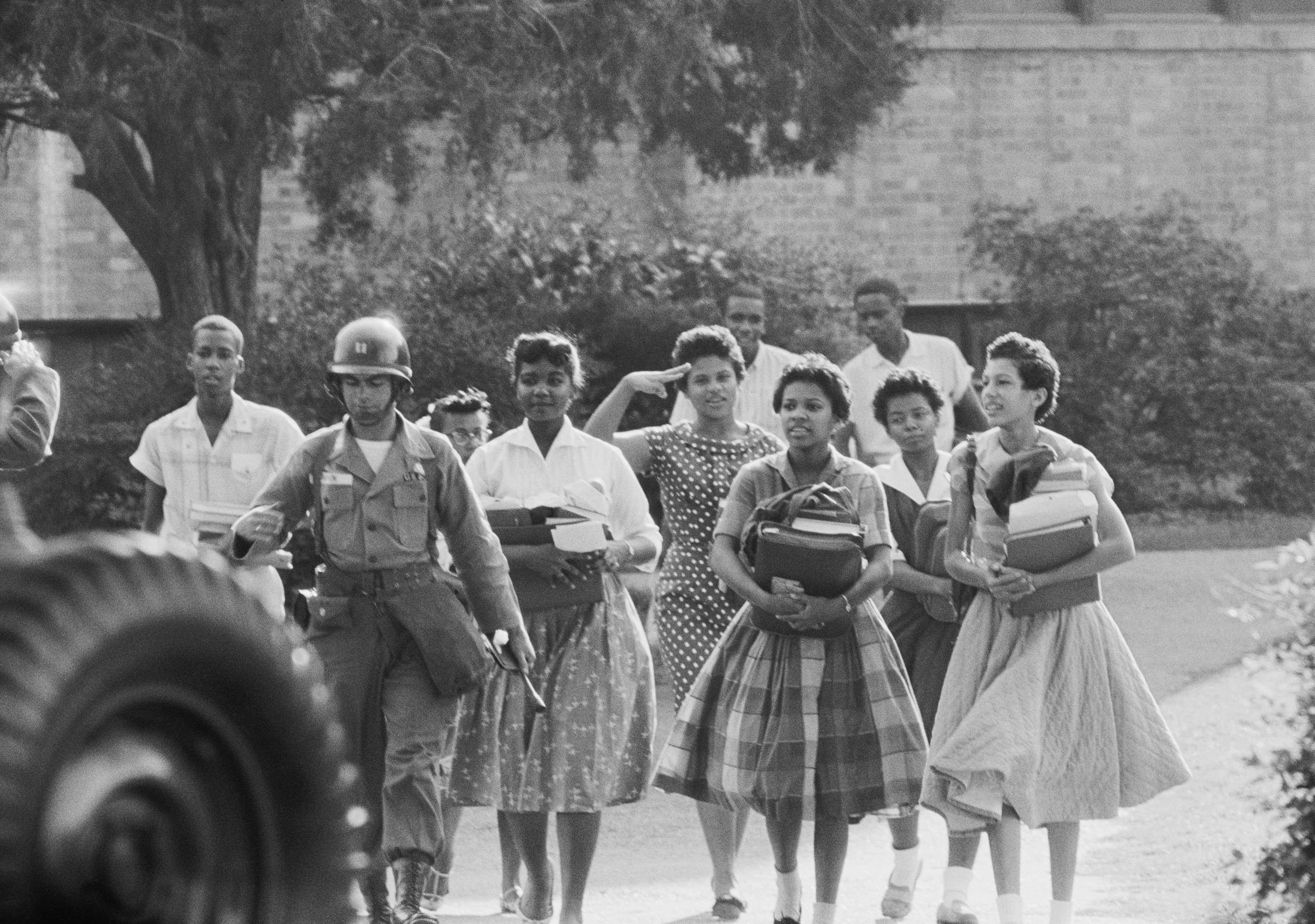 A black and white photo nine students walk from Little Rock's Central High School. The students are holding thier books while they follow a shoulder.