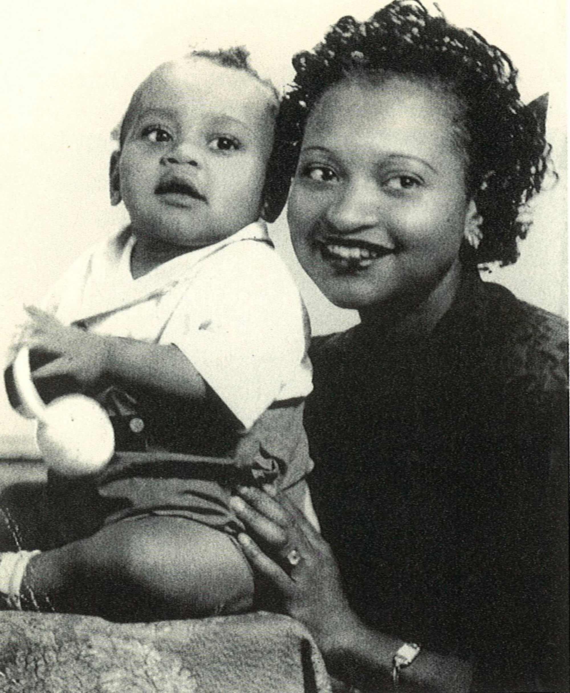 A black and white photograph of a smiling Mamie Till holding her baby, Emmett Till, as he plays with a rattle.