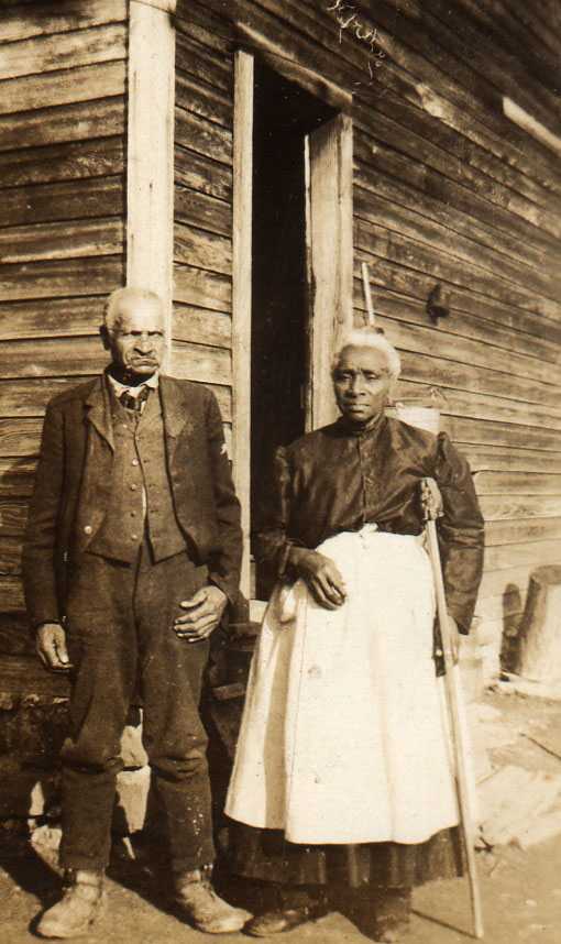 Two older people stand next to each other outside of a log cabin. On the left, the man is wearing a suit. On the right, the woman is a white apron.