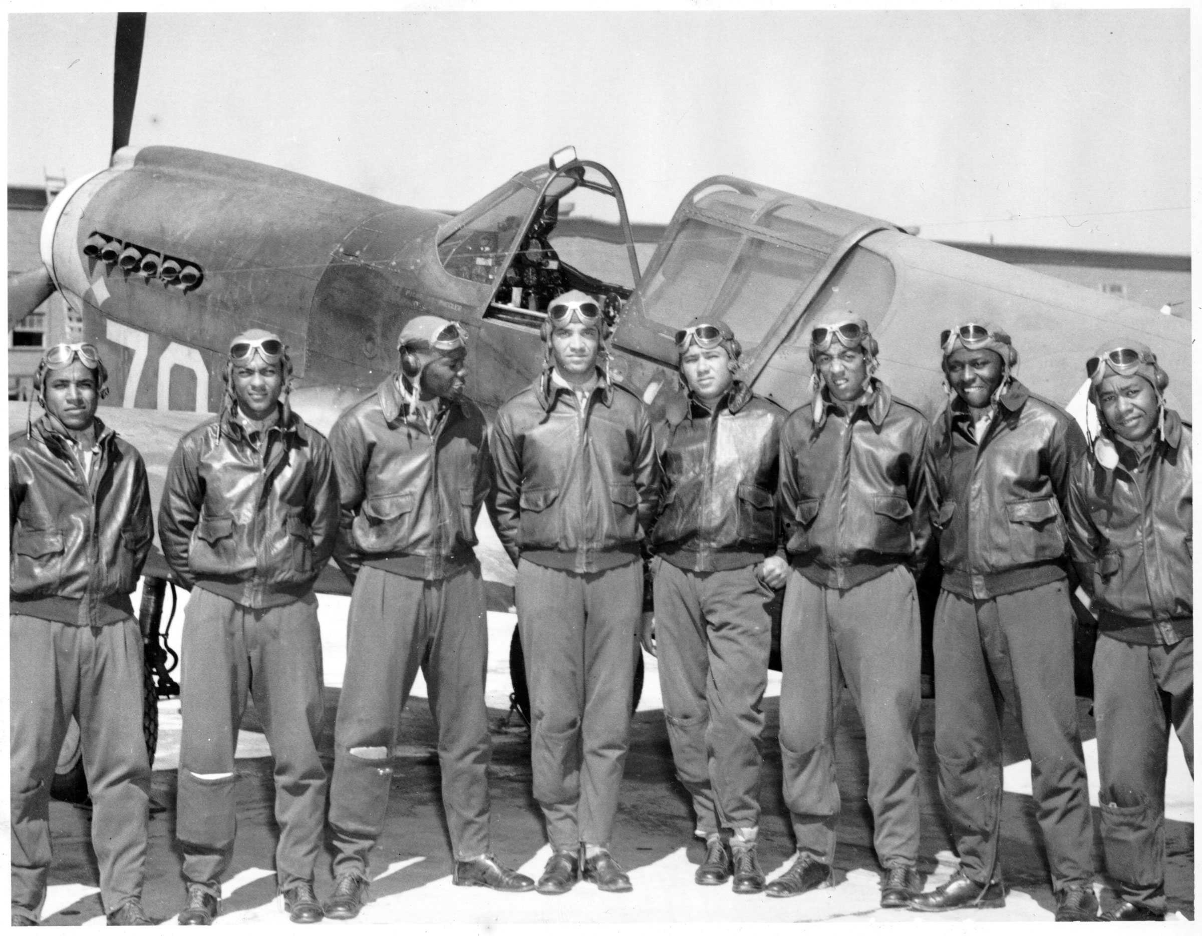 Black and white photograph showing a group of African American airmen posed before a military plane.  They all wear goggles and flight jackets.