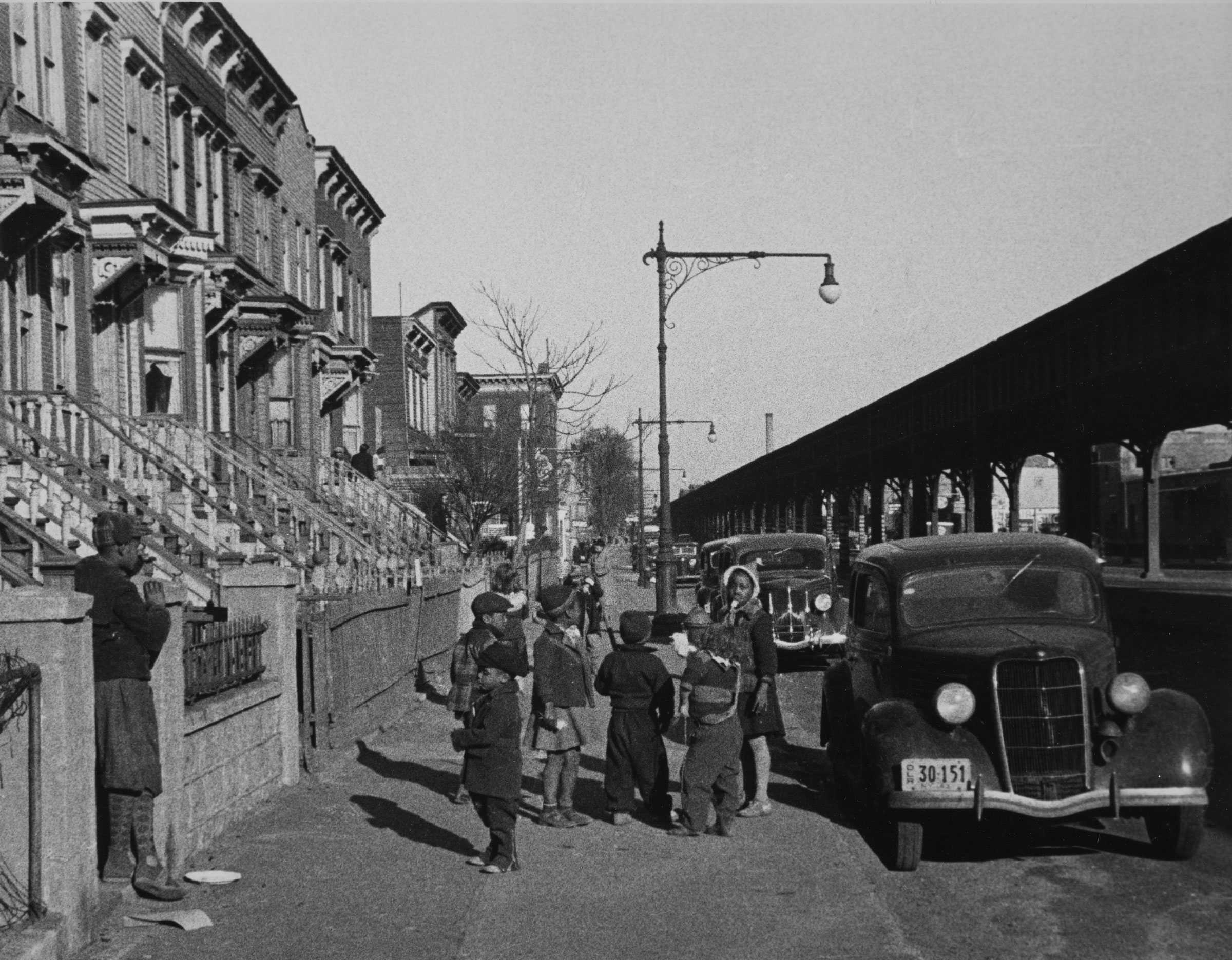 A black-and-white photograph of children loitering on a sidewalk. On the left, elevated trolley tracks.