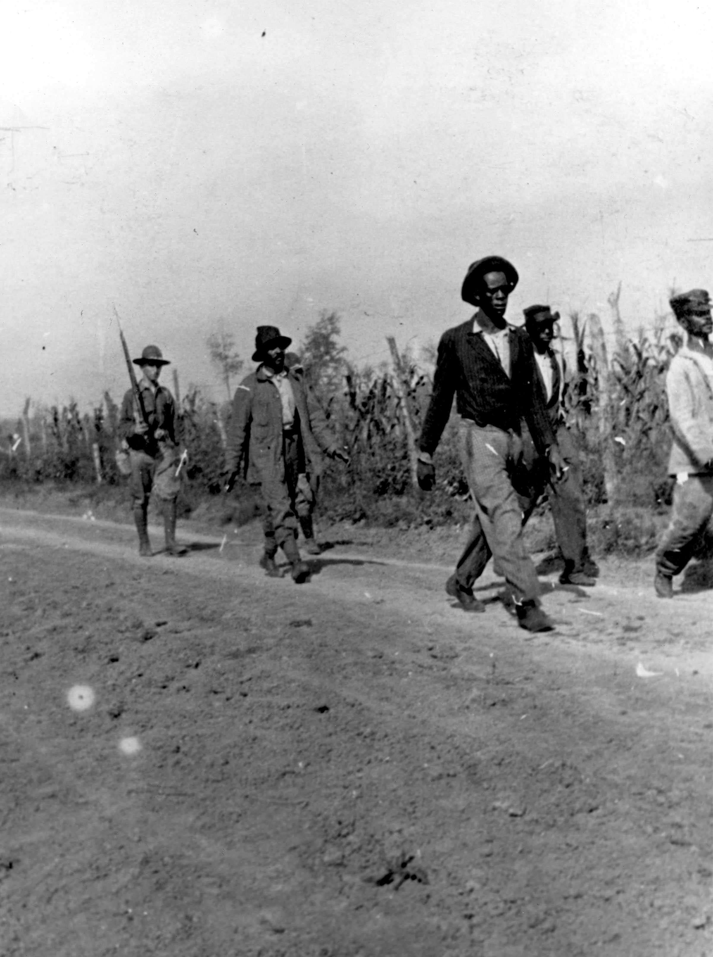 Soldier marching behind group of Black Men on a dirt road.