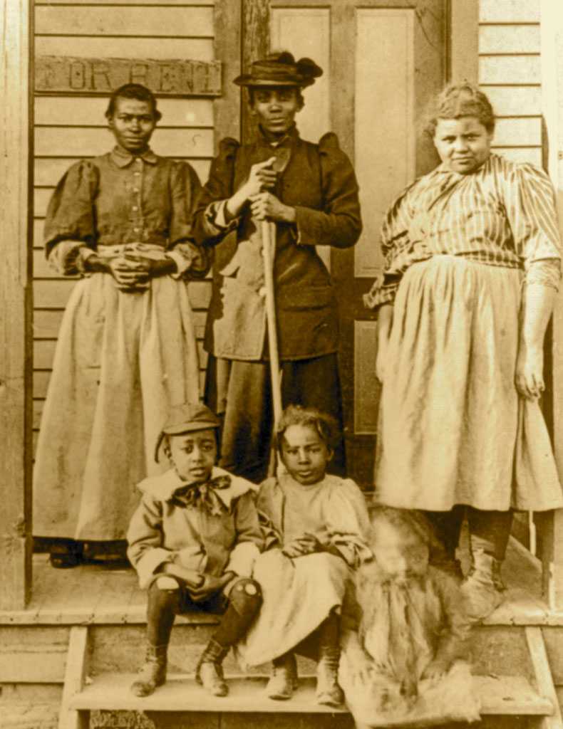 Sepia toned photograph of 3 women standing on a porch of a house with "FOR RENT" sign displayed on shingle.  There are three children seated on the steps below the women.