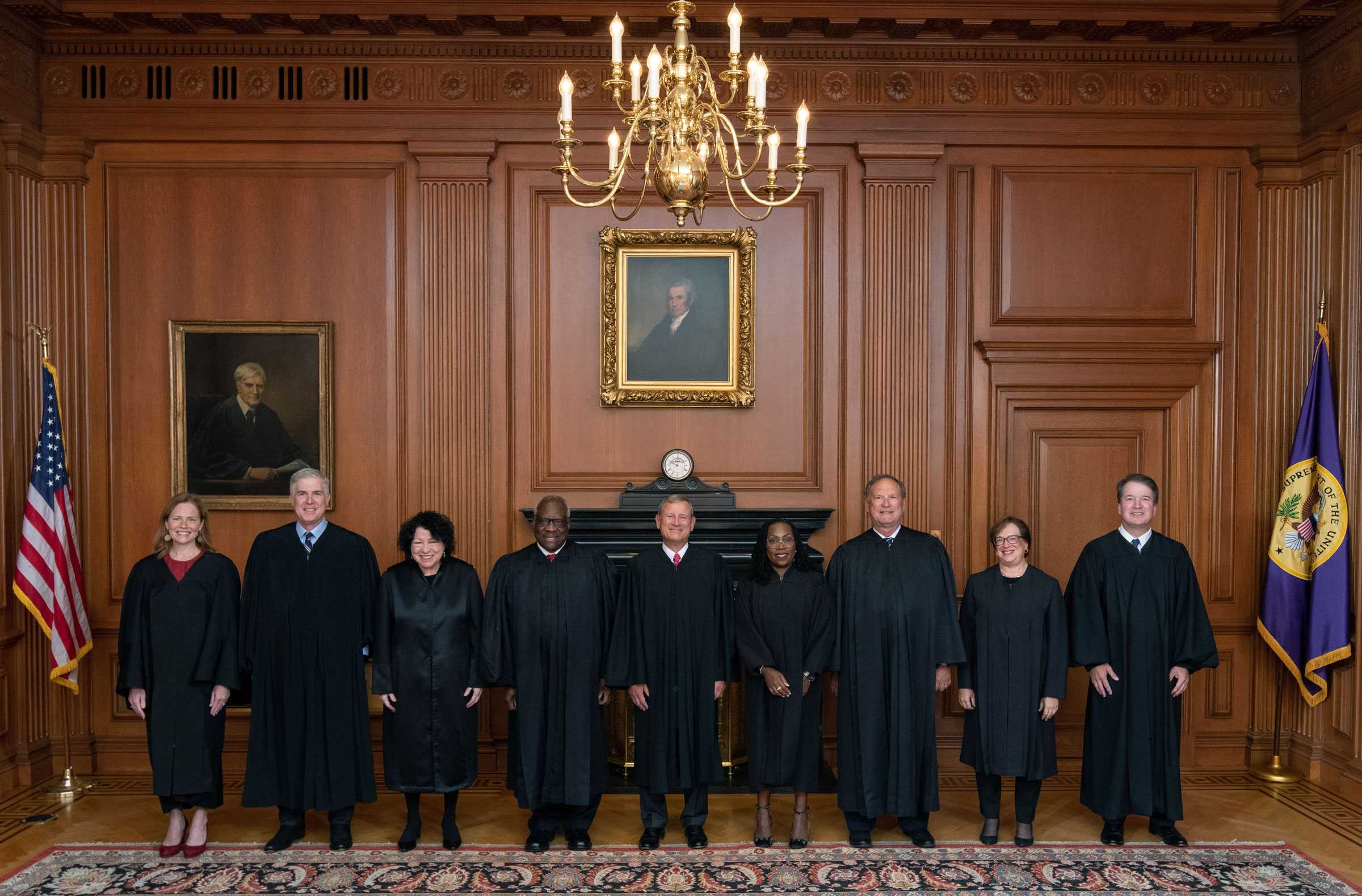 9 Supreme Court Justices stand together smiling and posing for a portrait inside the Supreme Court's building.