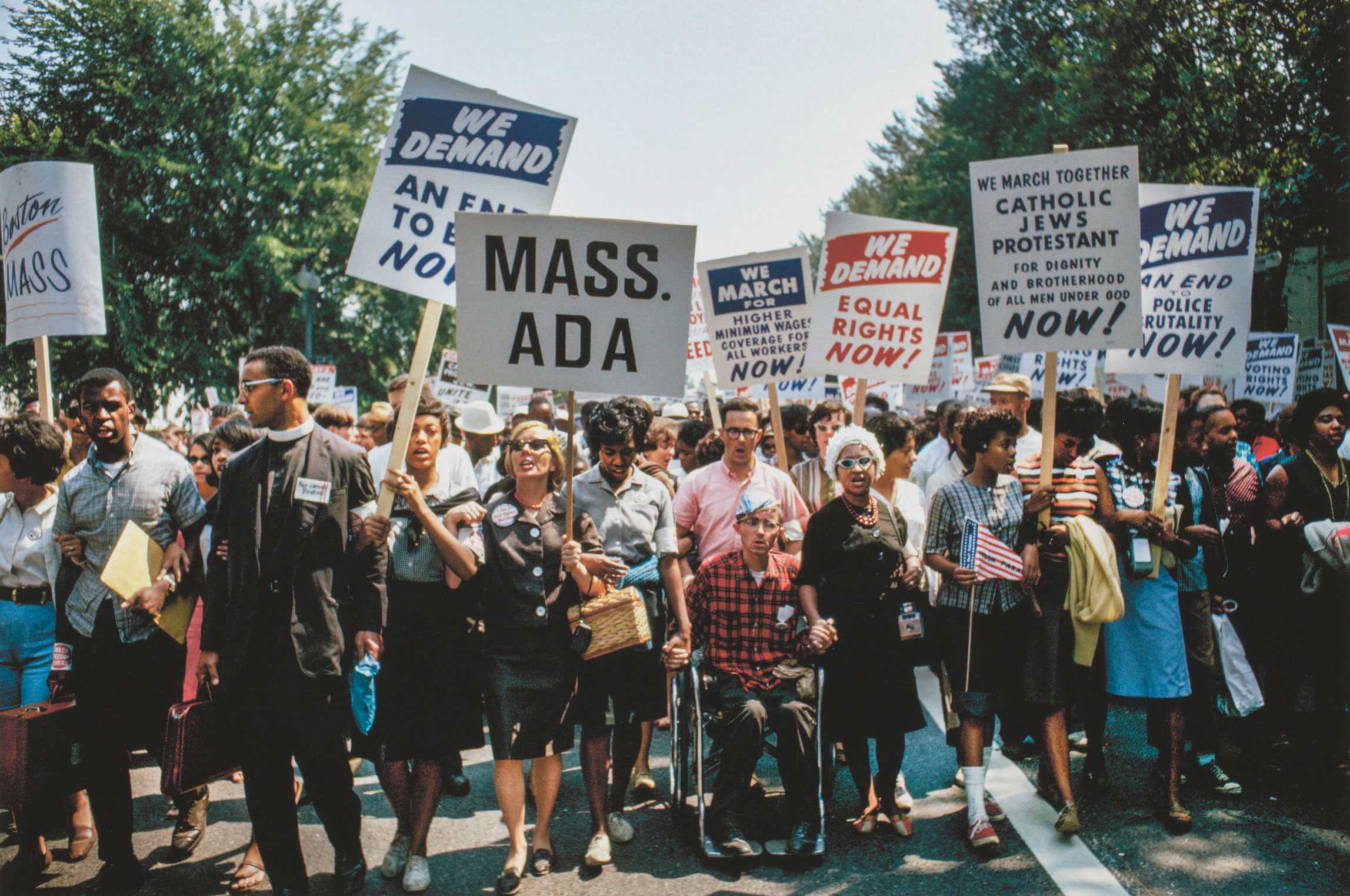 Color photograph of the 1963 March on Washington, showing a diverse group of protesters, including men and women, linking arms and holding signs advocating for equal rights.