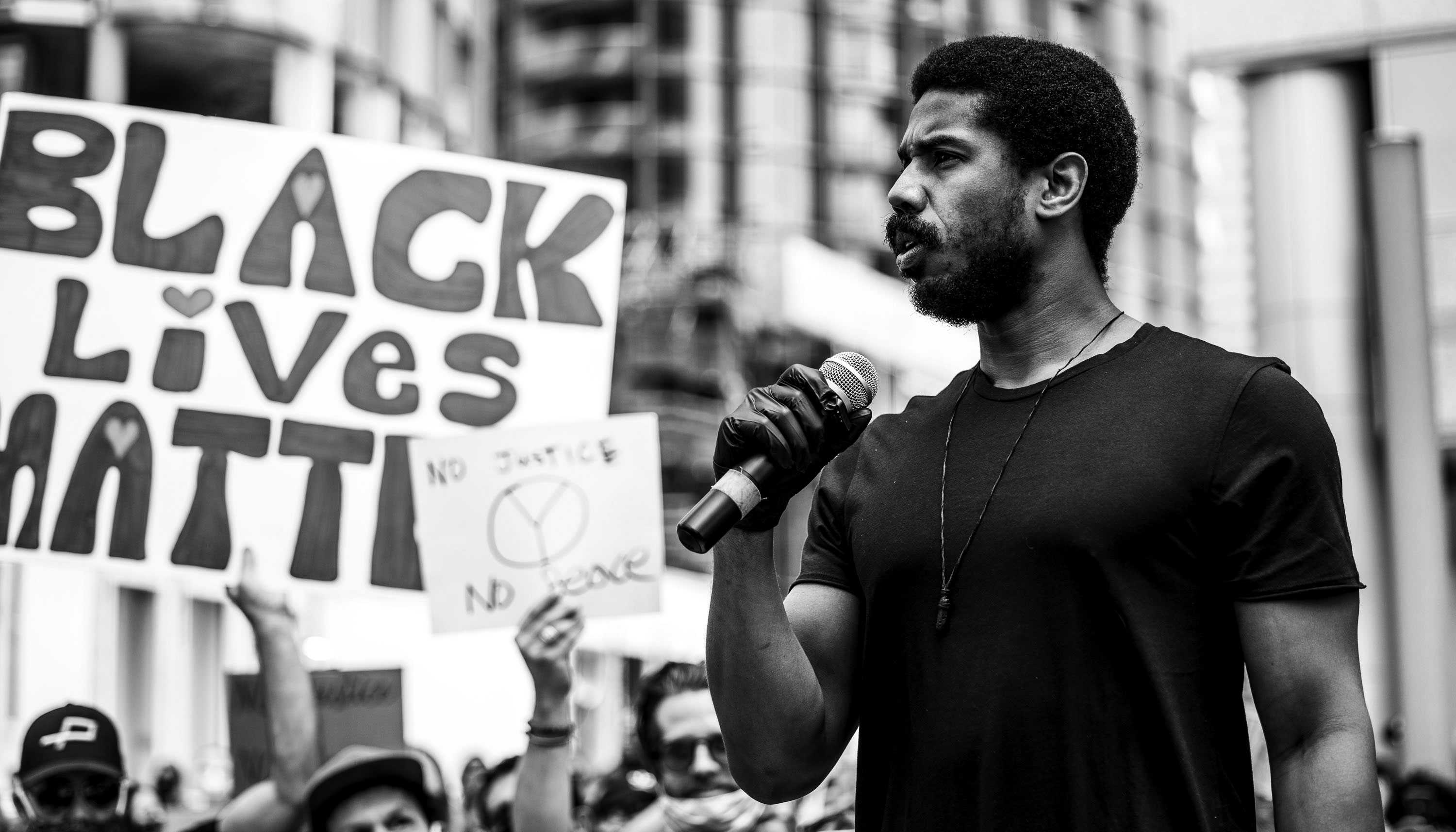 Michael B. Jordan speaking at a Black Lives Matter protest. He is shown from the waist up and turned in three quarters pose.