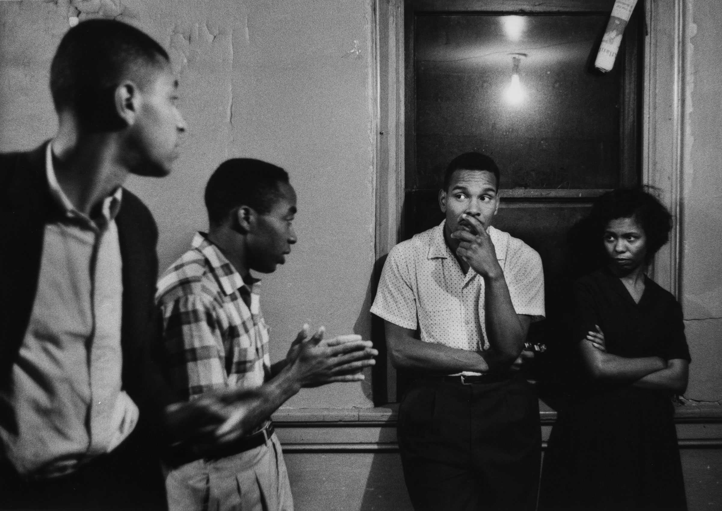 A black and white photograph of 4 people talking. Two lean against the window while the other two stand towards the center of the room.