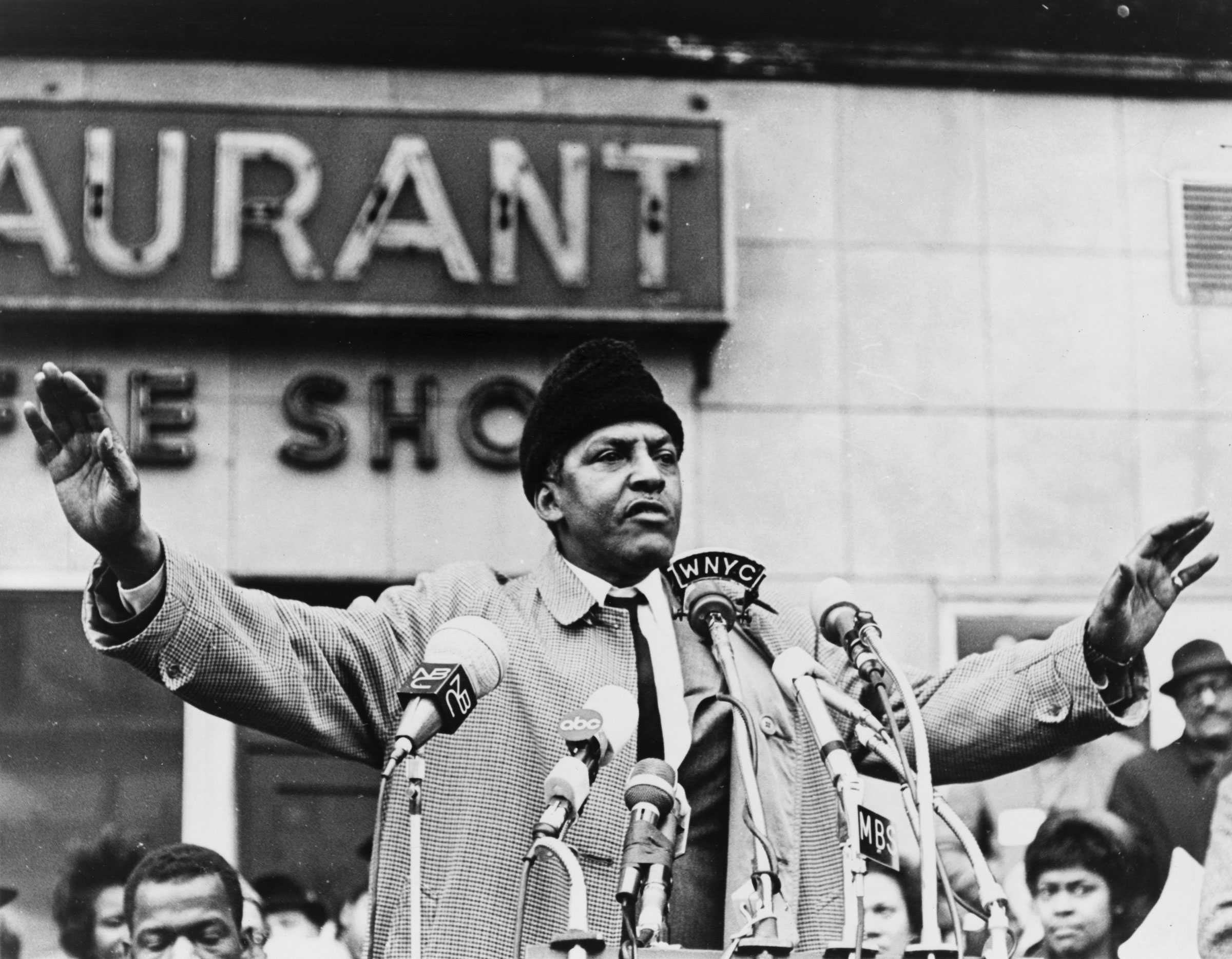 Bayard Rustin speaks into a group of microphones. Dressed in a coat and hat, his hands are stretched out to his sides to emphasize his points.