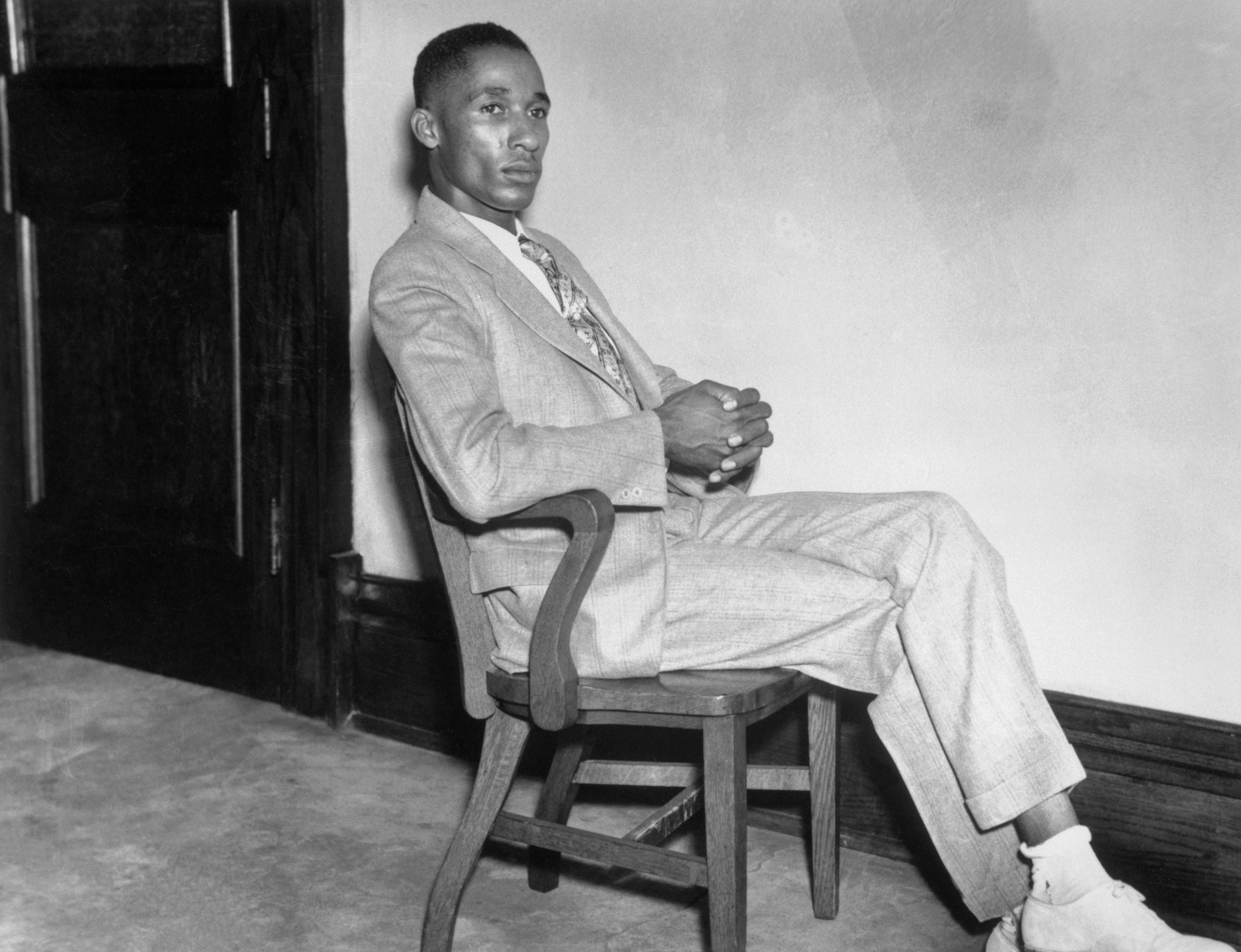 Black and white photograph of Black man seated in a wooden chair with crossed legs and hands folded in lap.  He is wearing a light colored suit and patterned neck tie.