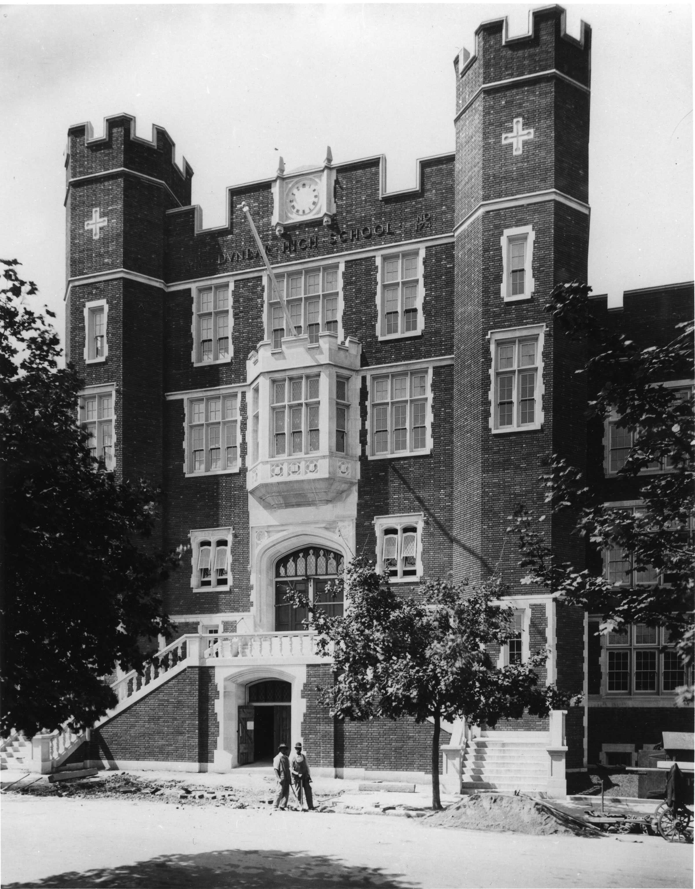 Black and white image of a brick building with white  trim. The building has two towers each with a white cross on them.  There is a clock between the two towers with the words "DUNBAR HIGH SCHOOL" below.