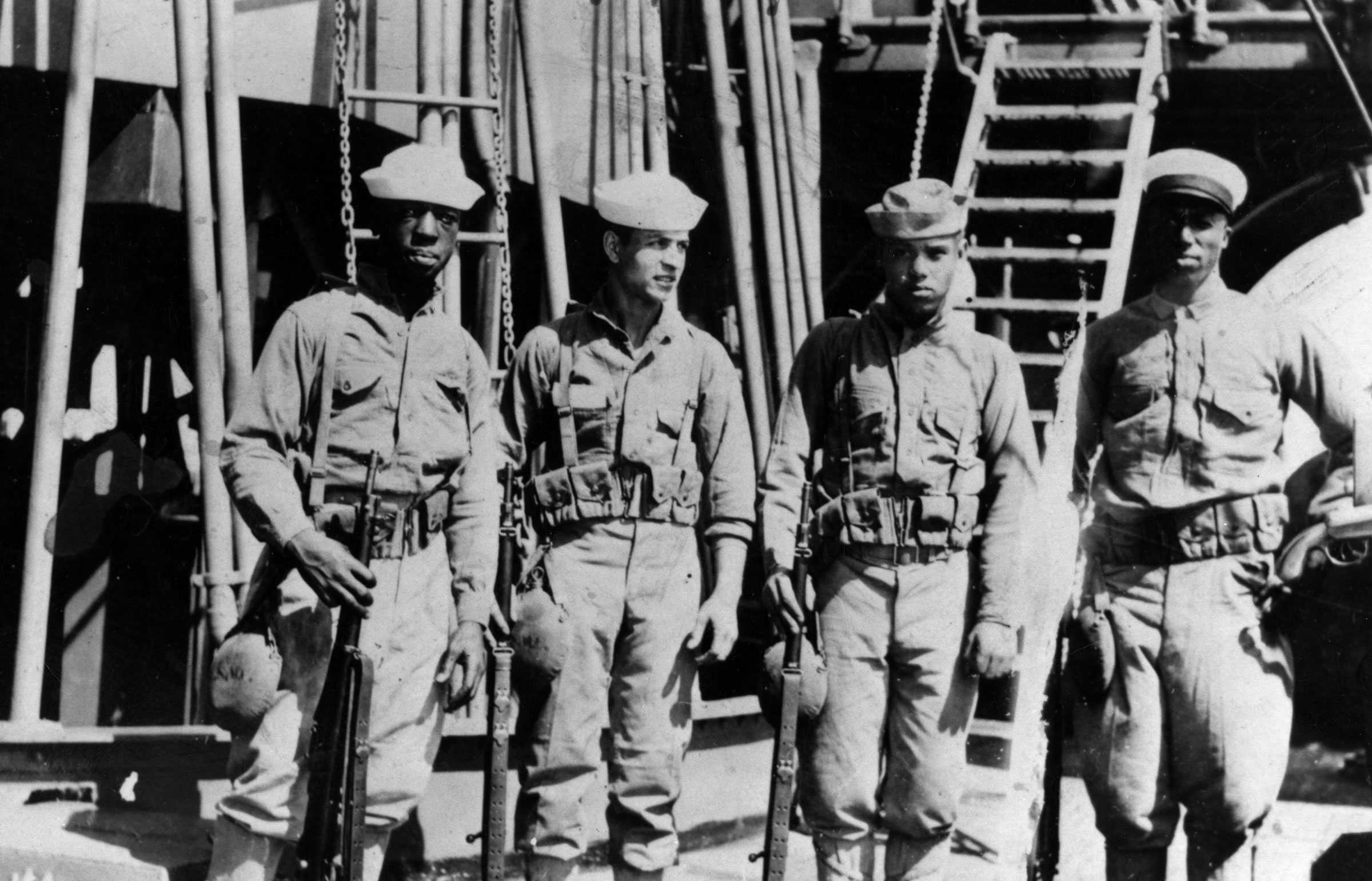 Black and white photograph of 4 sailors with guns and ammunition belts standing before a ship.