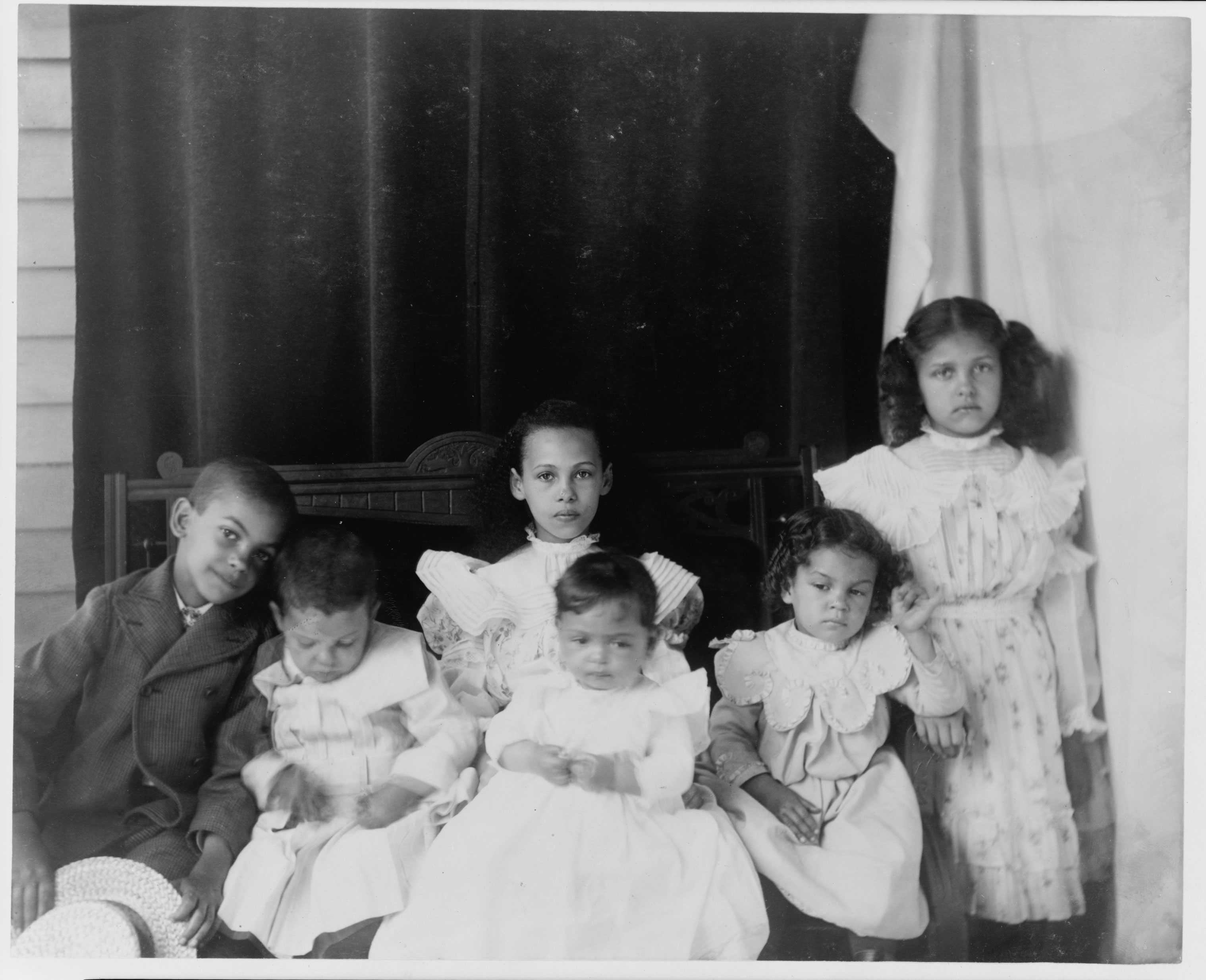 6 children, ranging in the ages of a baby to about 8 years old, sit together in white dresses and formal wear.