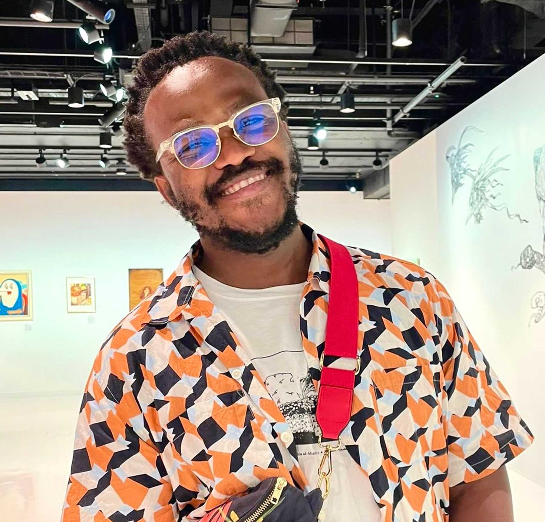 Illustrator Pola Maneli is smiling while he pose for a picture in an art gallery. He is wearing a black and orange geometric shirt.