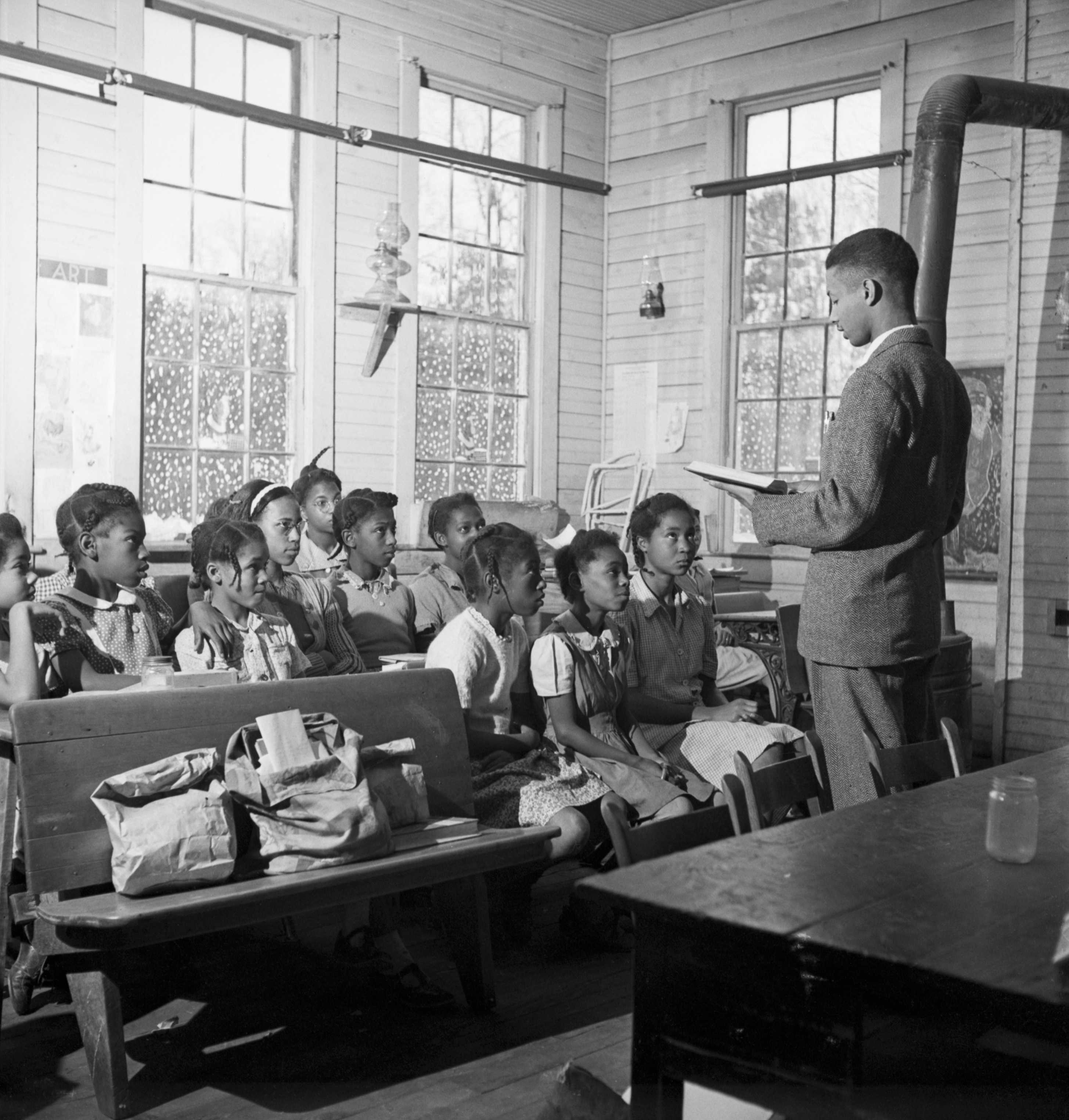 Black and white photograph of children seated in a classroom on benches.  There is also a male teacher reading to them from an open book.