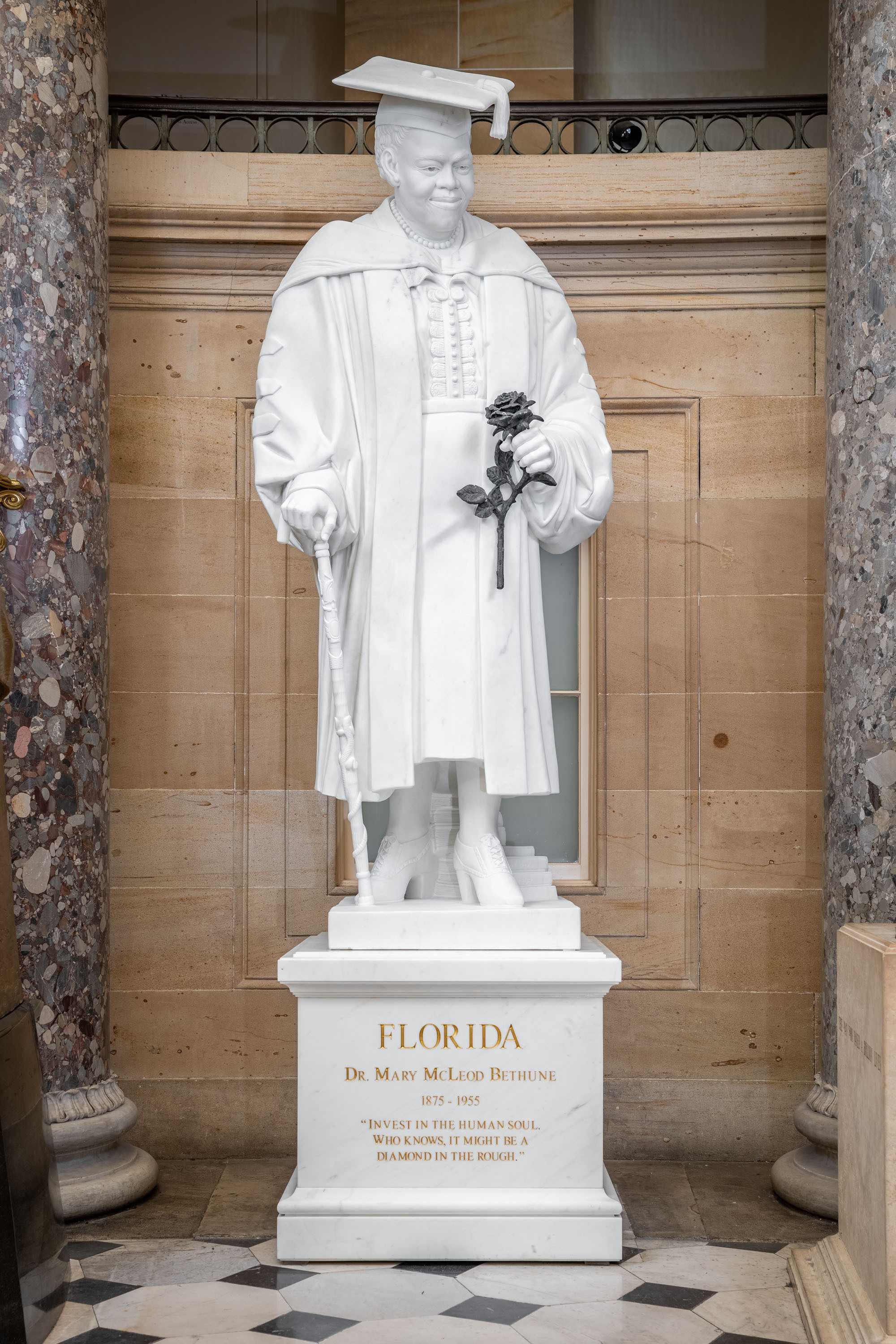 Color photograph of marble statue of woman dressed in academic regalia holding a black rose and walking cane.  The statue is on top of a marble pedestal engraved with "FLORIDA / DR. MARY MCLEOD BETHUNE / 1875-1955 / "INVEST IN THE HUMAN SOUL,. WHO KNOWS IT MIGHT BE A DIAMOND IN THE ROUGH."