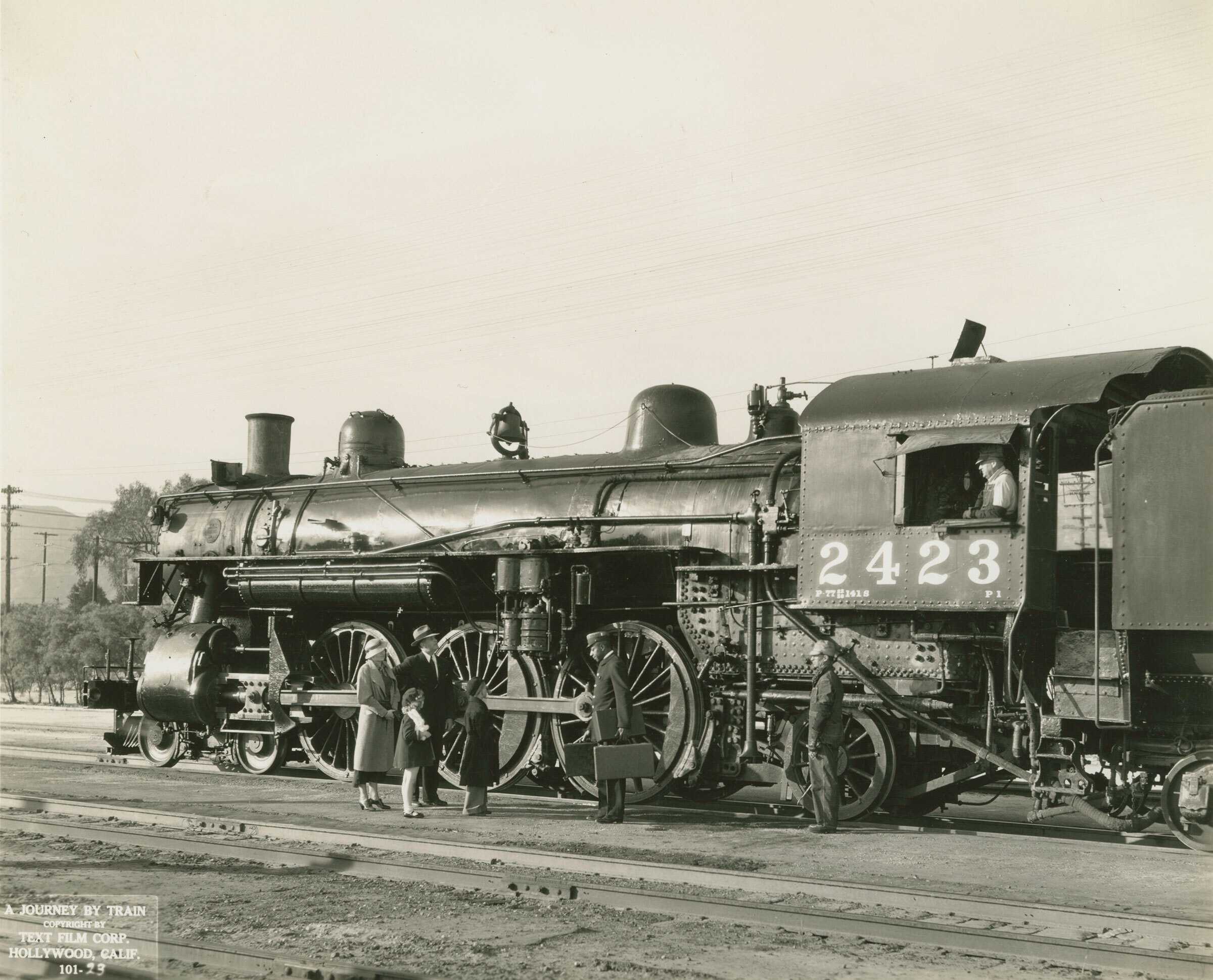 Black and white photograph depicting an outdoor scene of a train yard. Inside the train you can see a train engineer standing at the window.