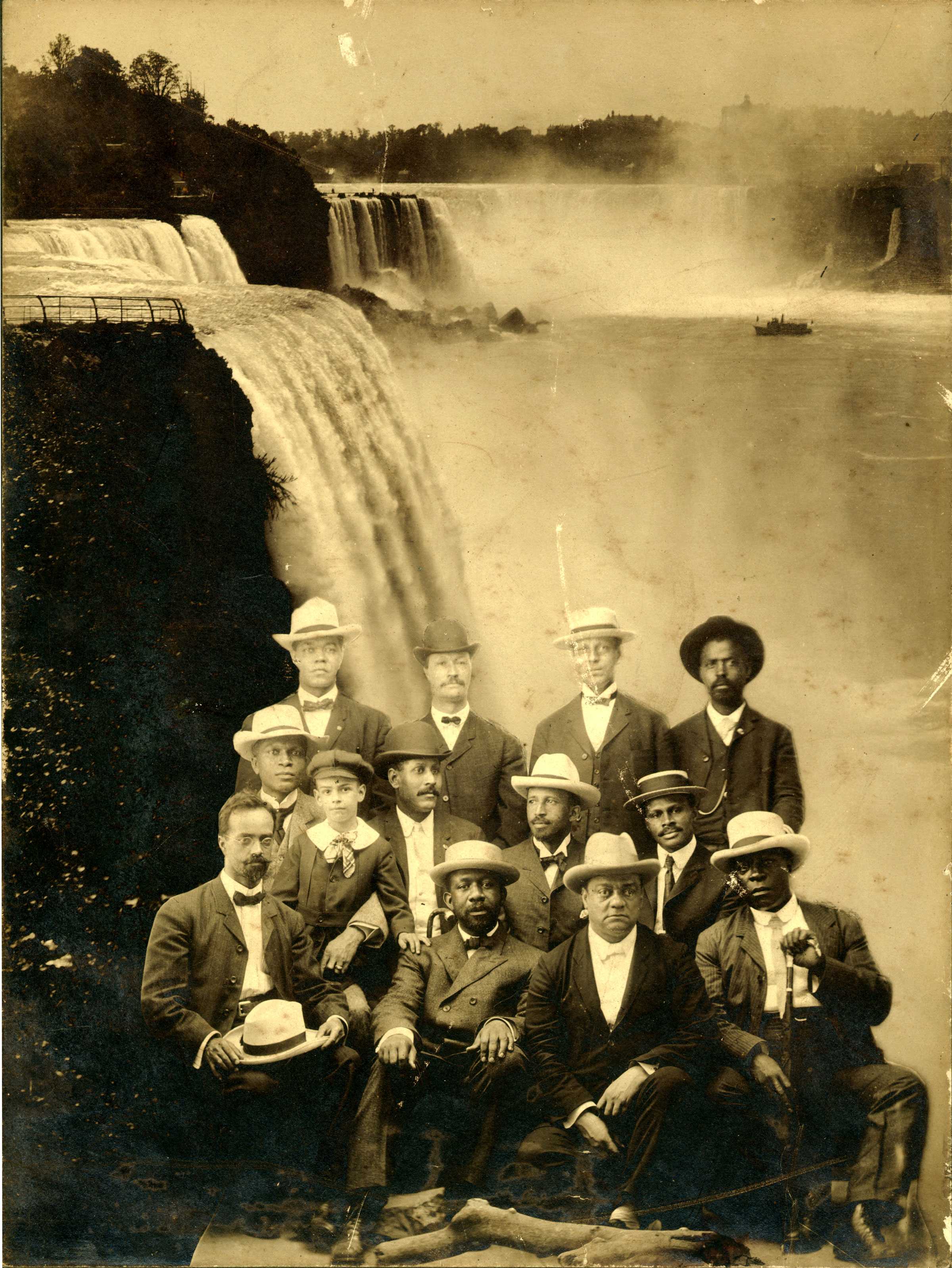 Black and white photograph showing a group of individuals sitting in front of cascading waterfall.   Trees surround the falls, and a boat appears near the mist.