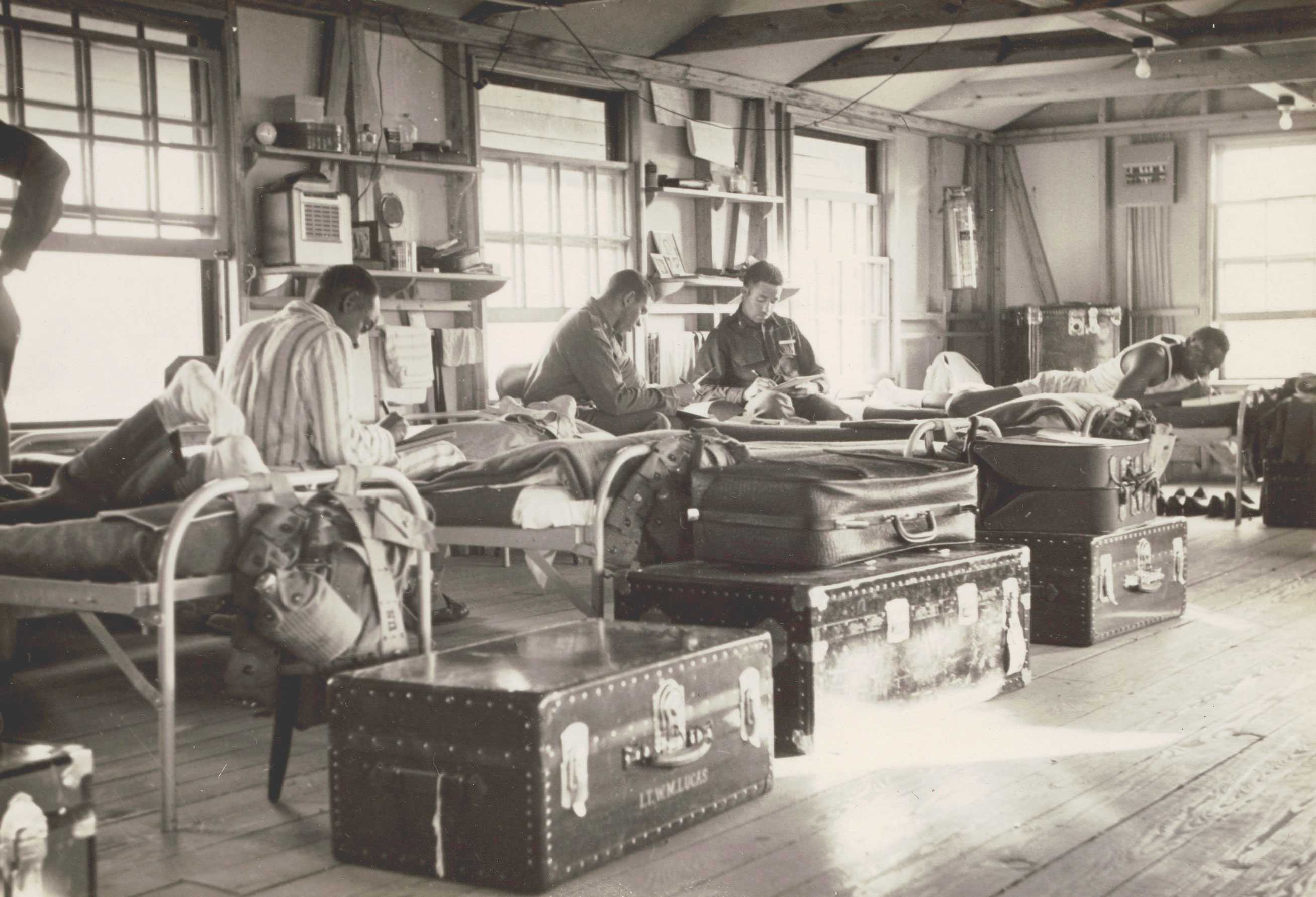 A black-and-white photo of a military bunkhouse with four men on beds writing letters and various items on shelves.