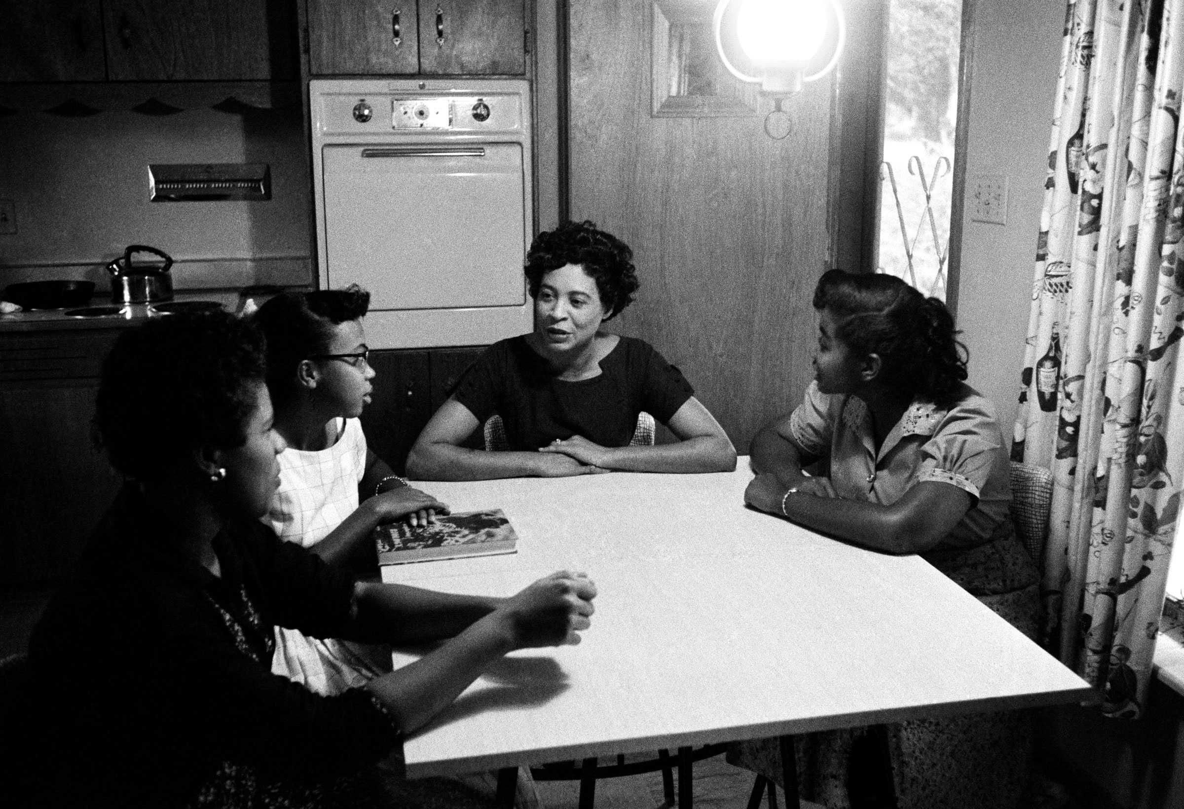 Daisy Bates sits and talks with 3 girls of the Little Rock 9 at a kitchen table. A single pendant above the lights the kitchen.