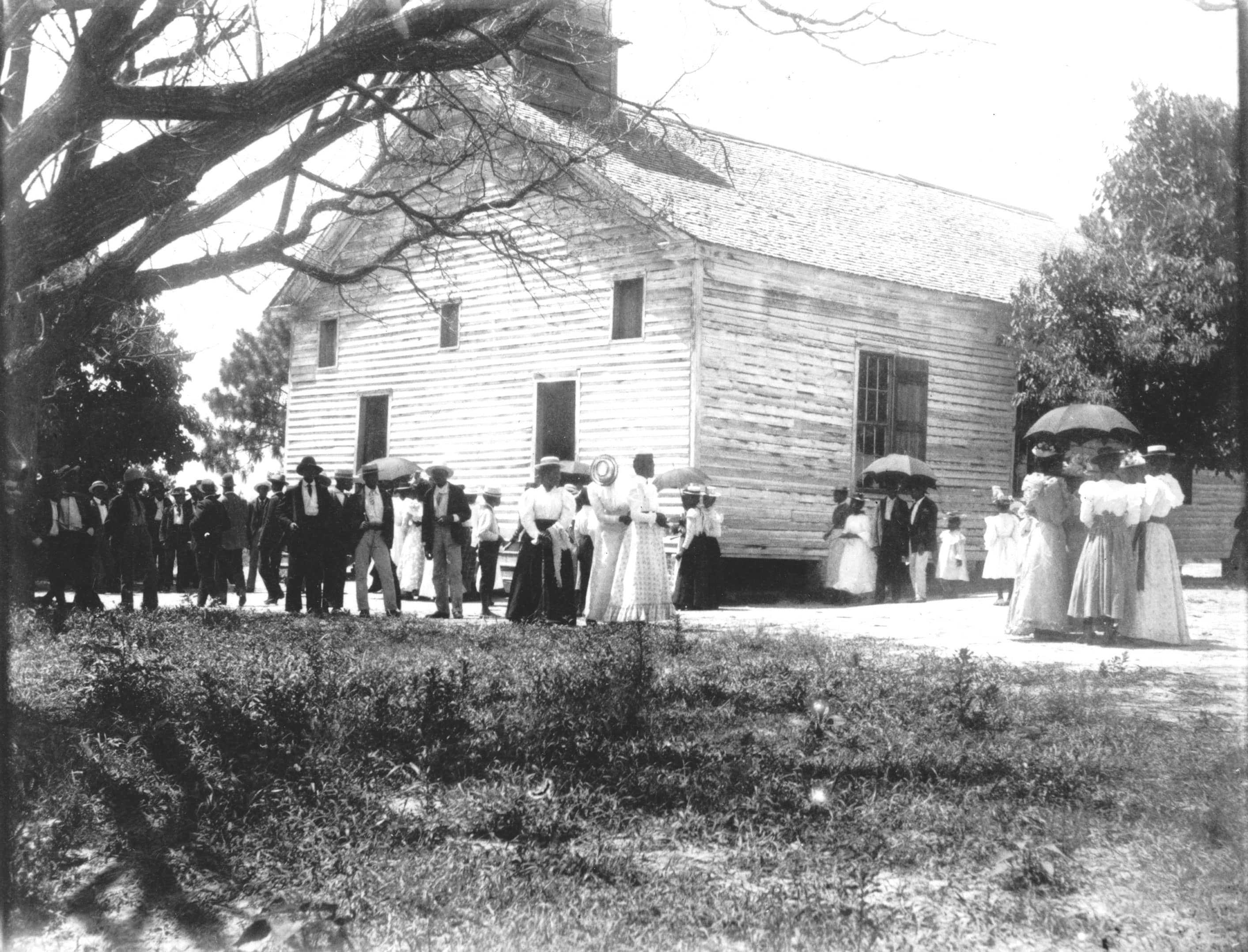 A large group of men and woman African American congregate outside of a white church.