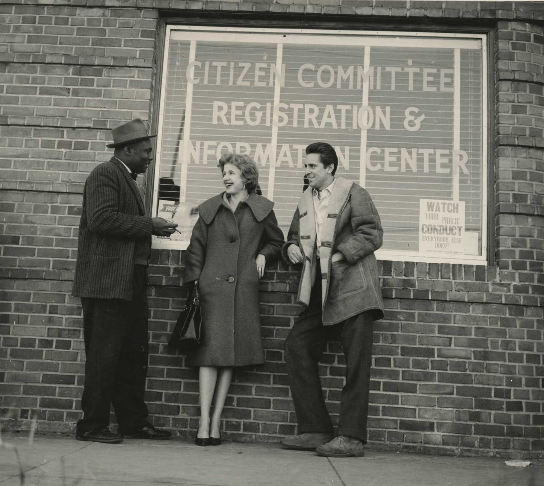 A black and white photograph of Esau Jenkins talking to Guy Carawan and a woman outside the window of the Citizen Committee Center