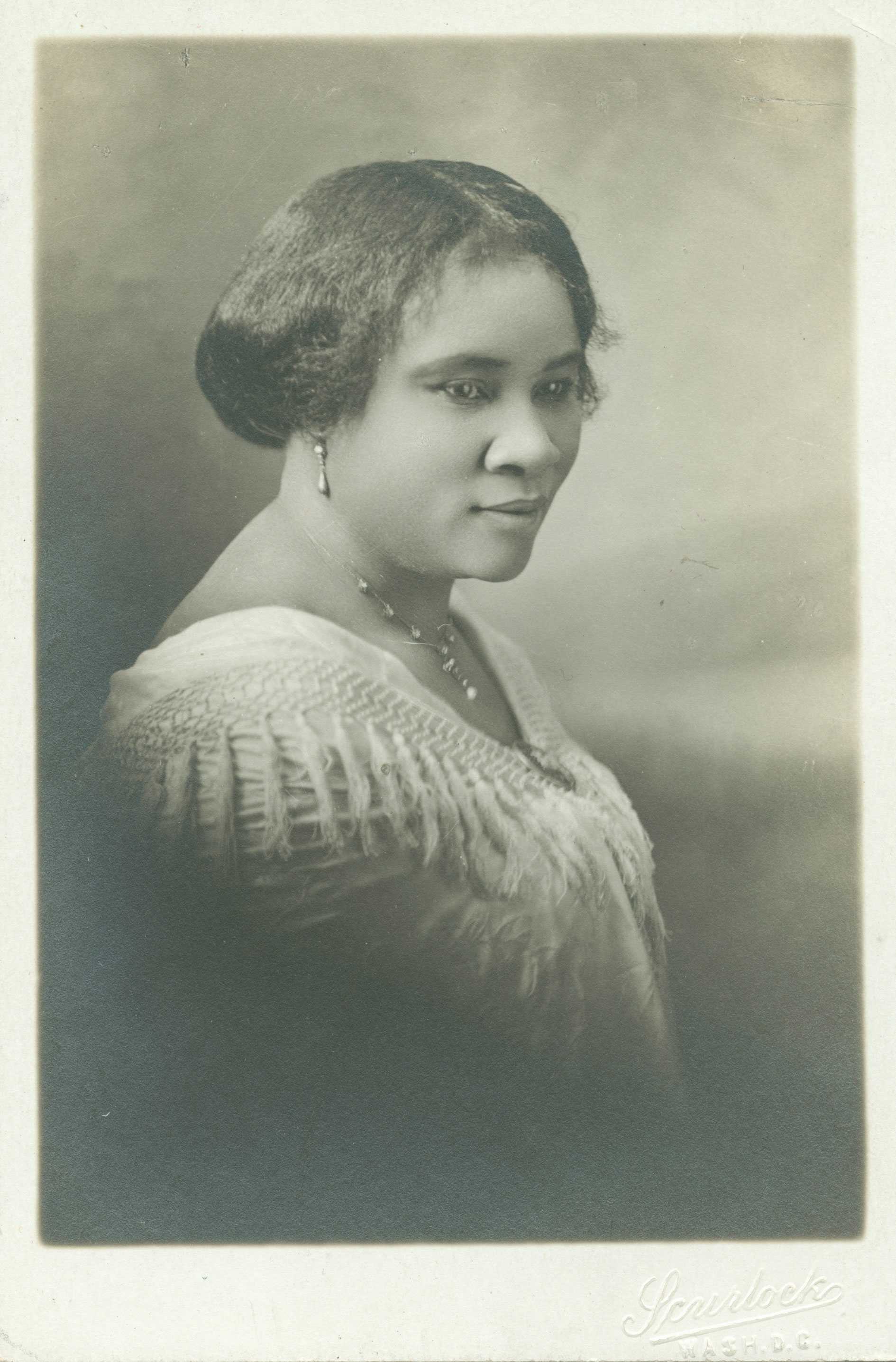 A black and white bust-length portrait of Madam C.J. Walker. She sits sideways, her head turned slightly right towards the viewer.