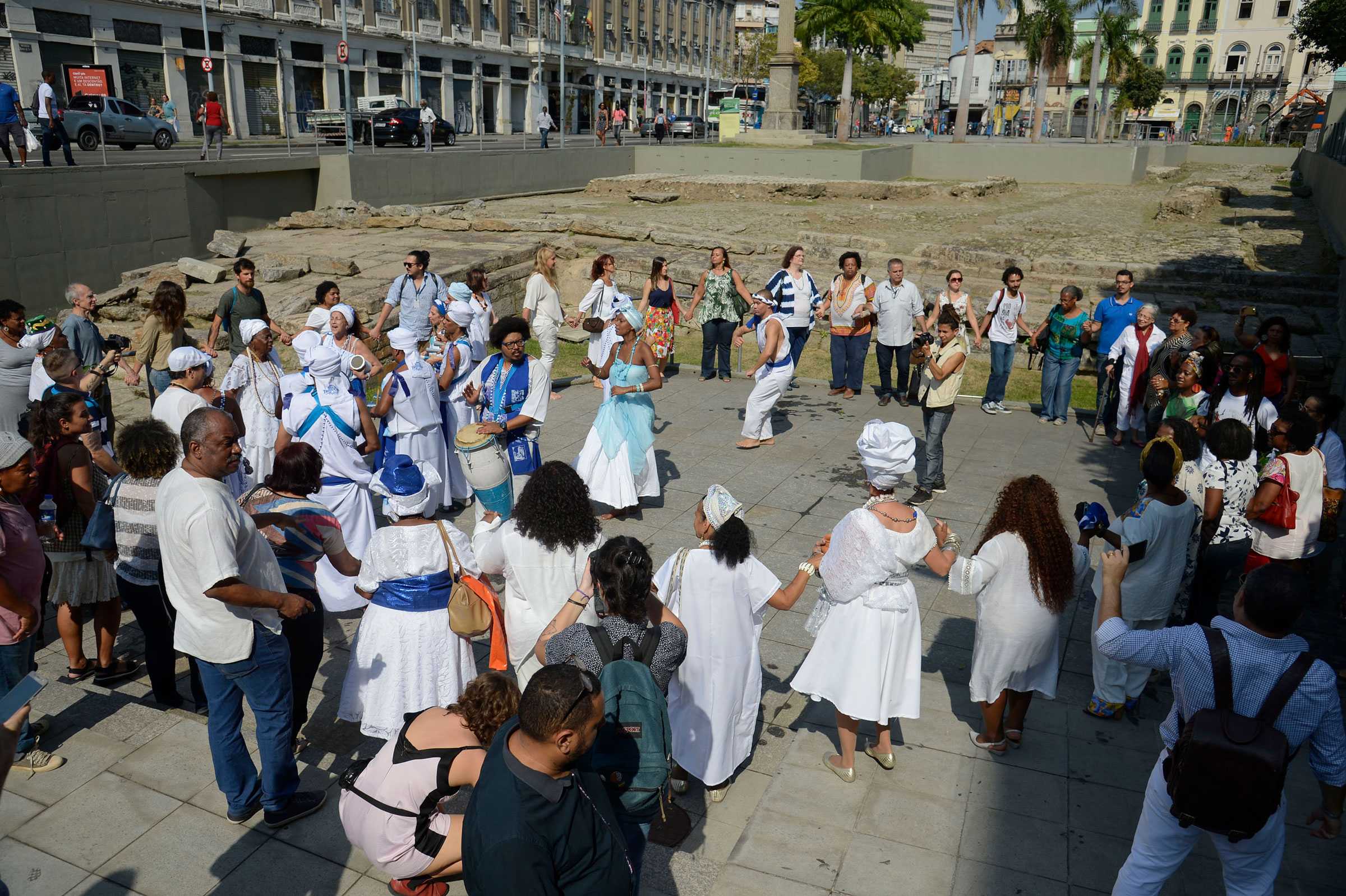 A group of people within Valongo Wharf, slightly below street level. Many are holding hands in a circle and dressed in white.