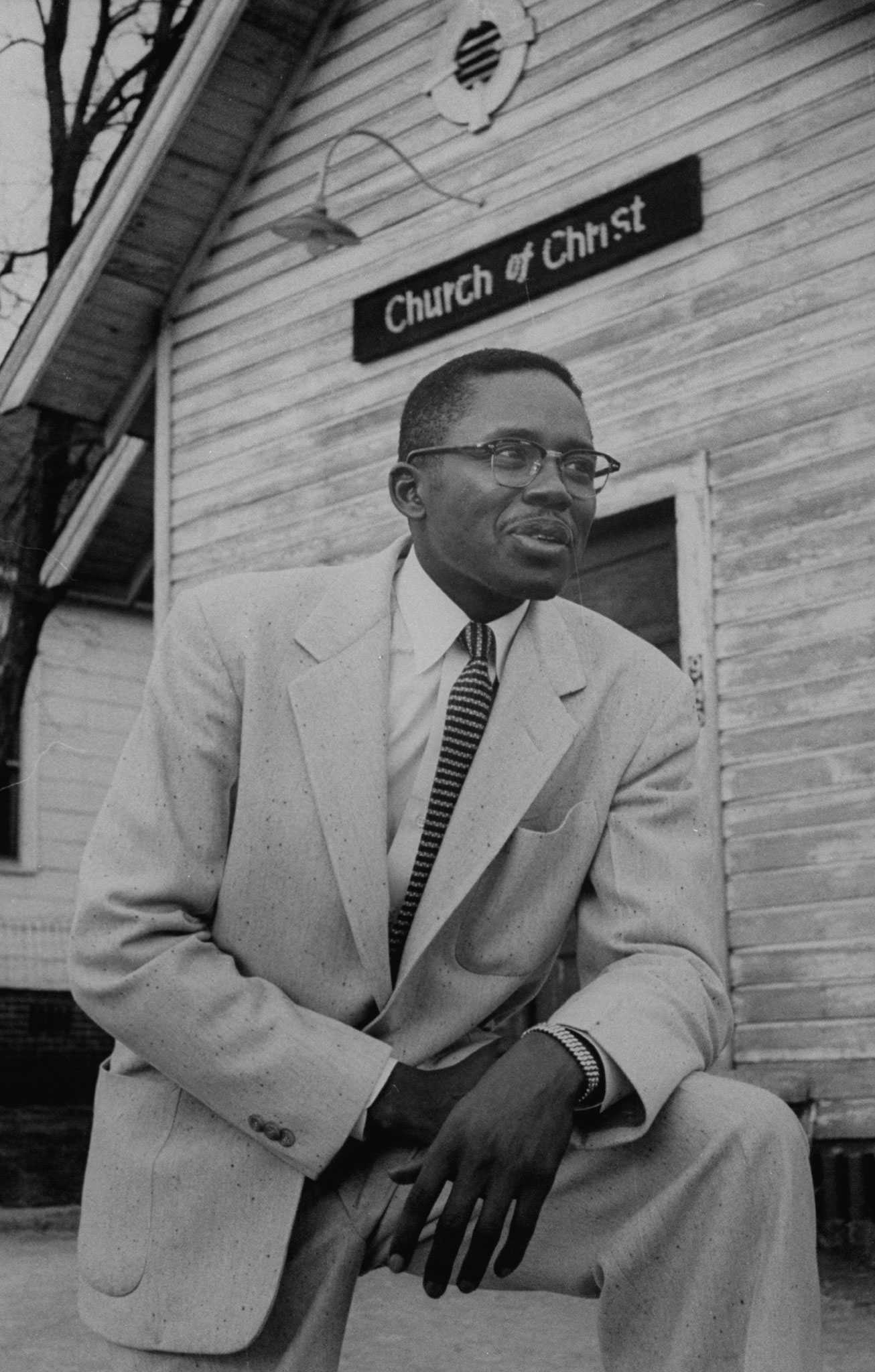 In a light colored suit and striped tie, Fred Gray, looking off camera, kneels in front of the Newton Church of Christ.