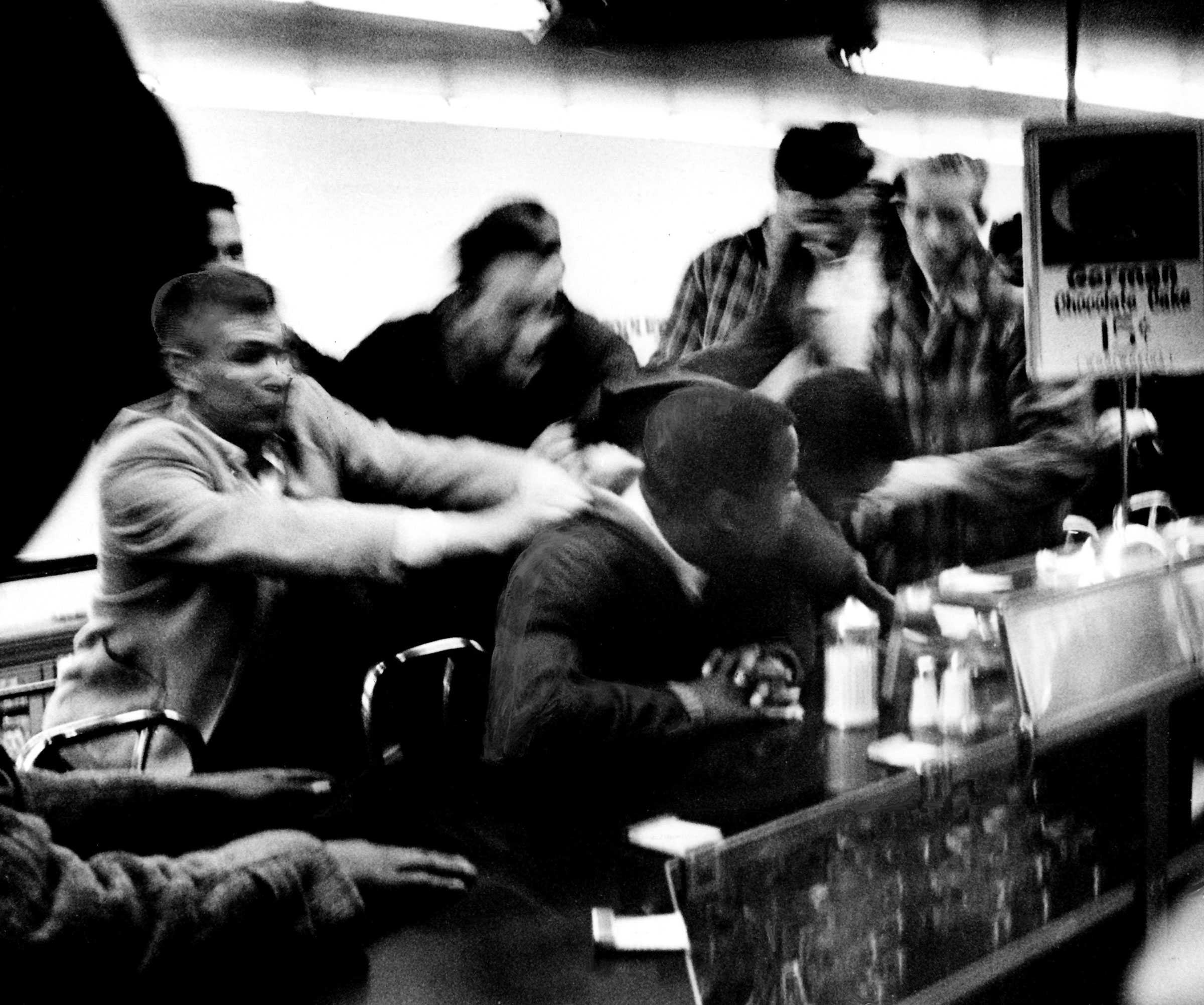 A black and white photograph of white men pulling Black protesters from the counter. The image is blurry from the men's quick movements.