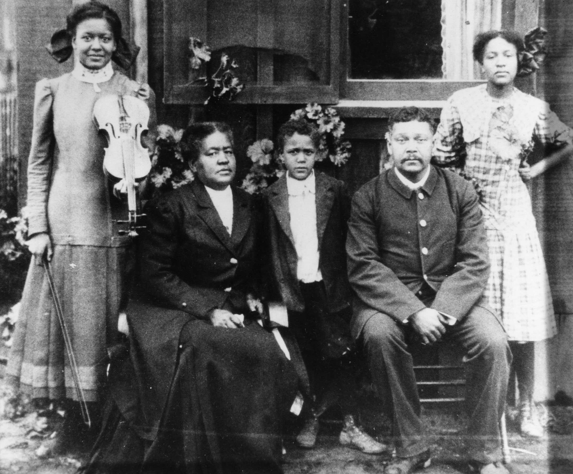 Black and white photograph of five people posed in front of a brick-facade building. 3 are seated in the front row, while two young women stand on either side.  One of the young women holds a violin.