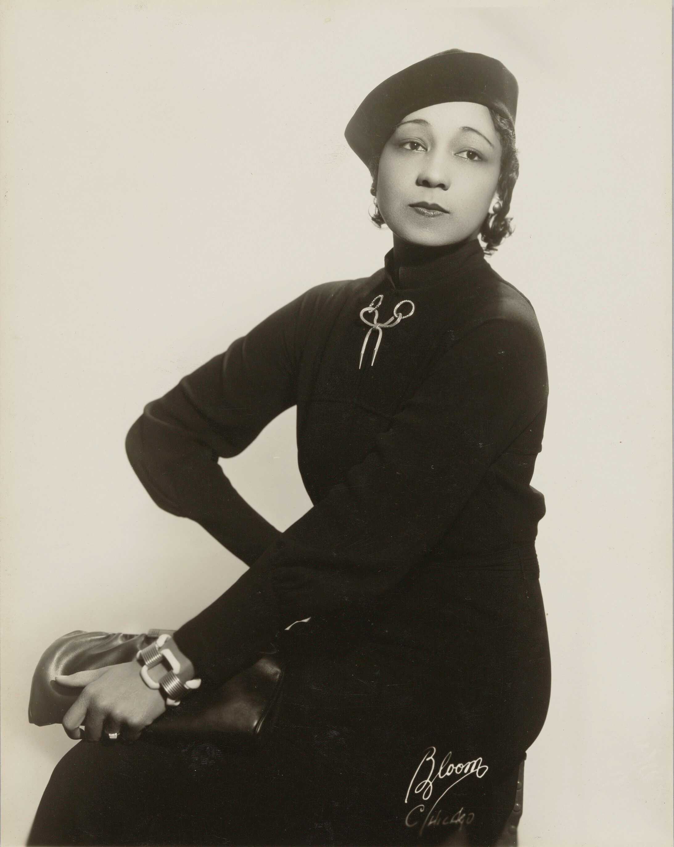 Black and white portait of African American woman seated.  She wears a dress with a high turtleneck and long sleeves. On her head is a black beret. At her neckline is a brooch in the shape of bow suspended from two rings, and on her left wrist is a large chunky chain link bracelet.