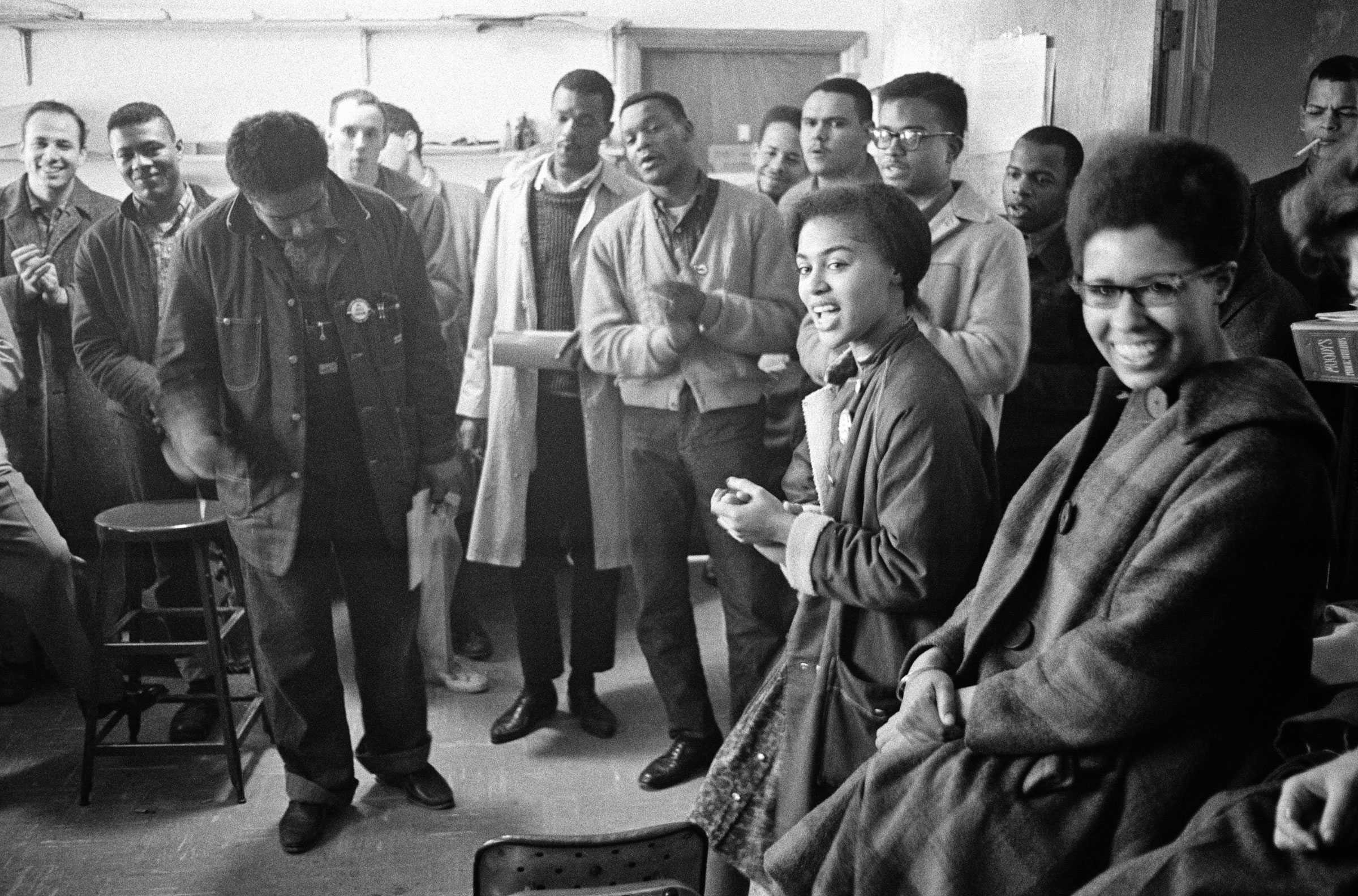 SNCC staff members gather together in the backroom in a semicircle. Some are laughing and smiling, while others listen.