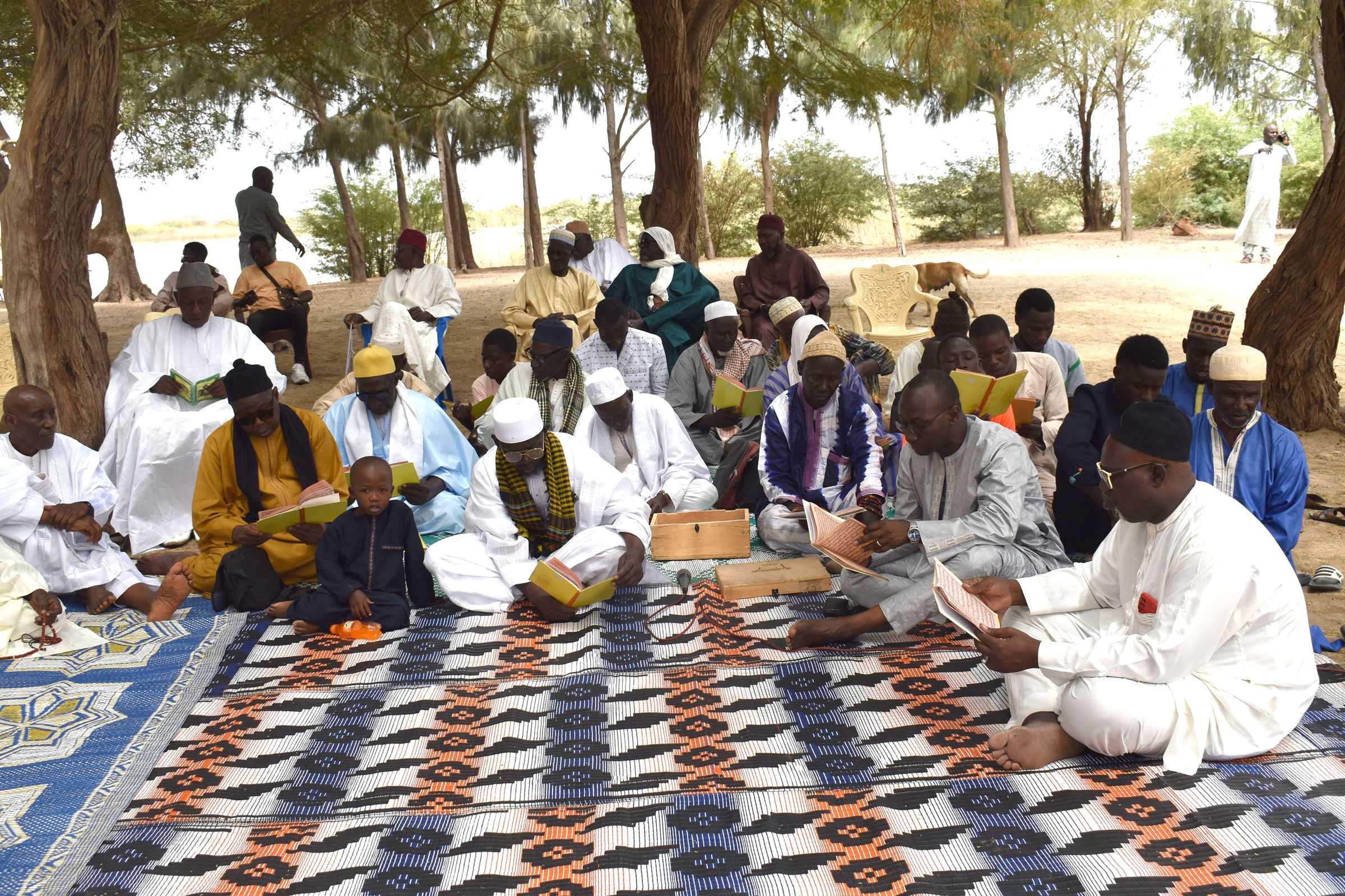 A group of men reading from the same yellow book. They sitting outside on a large blanket.