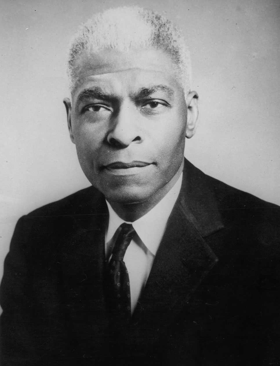 A black and white portrait of Benjamin May. He is wearing dark rimmed glasses and a dark suit.