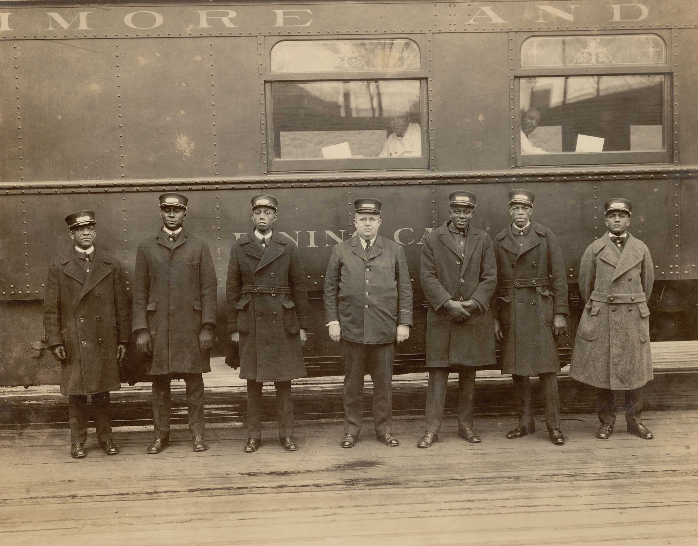 Sepia toned photograph of 7 Pullman Porters standing in front of a train car.  They are dressed in their uniforms.